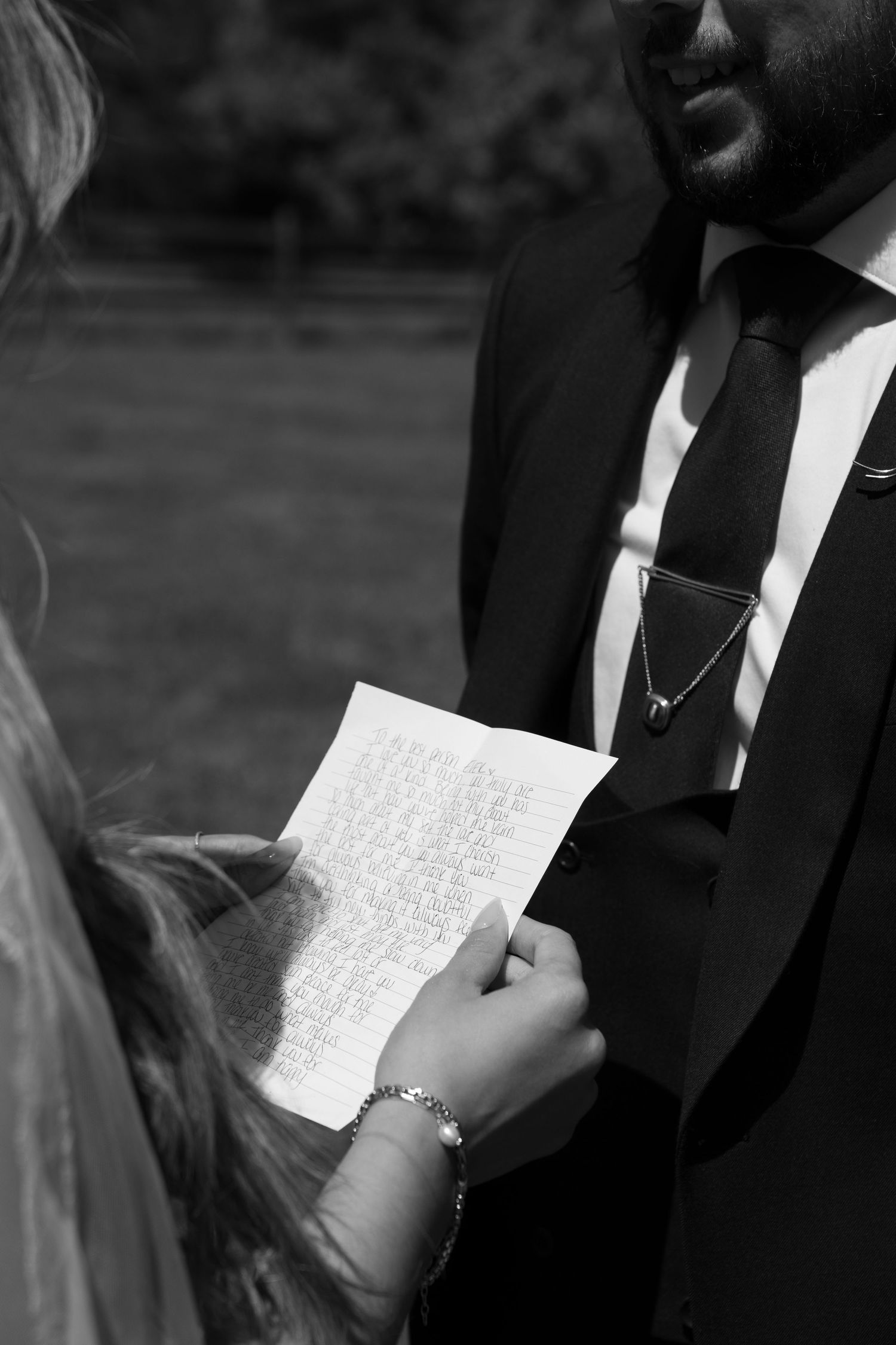 Black and white photo of bride reading handwritten wedding vows to groom in suit during outdoor ceremony.