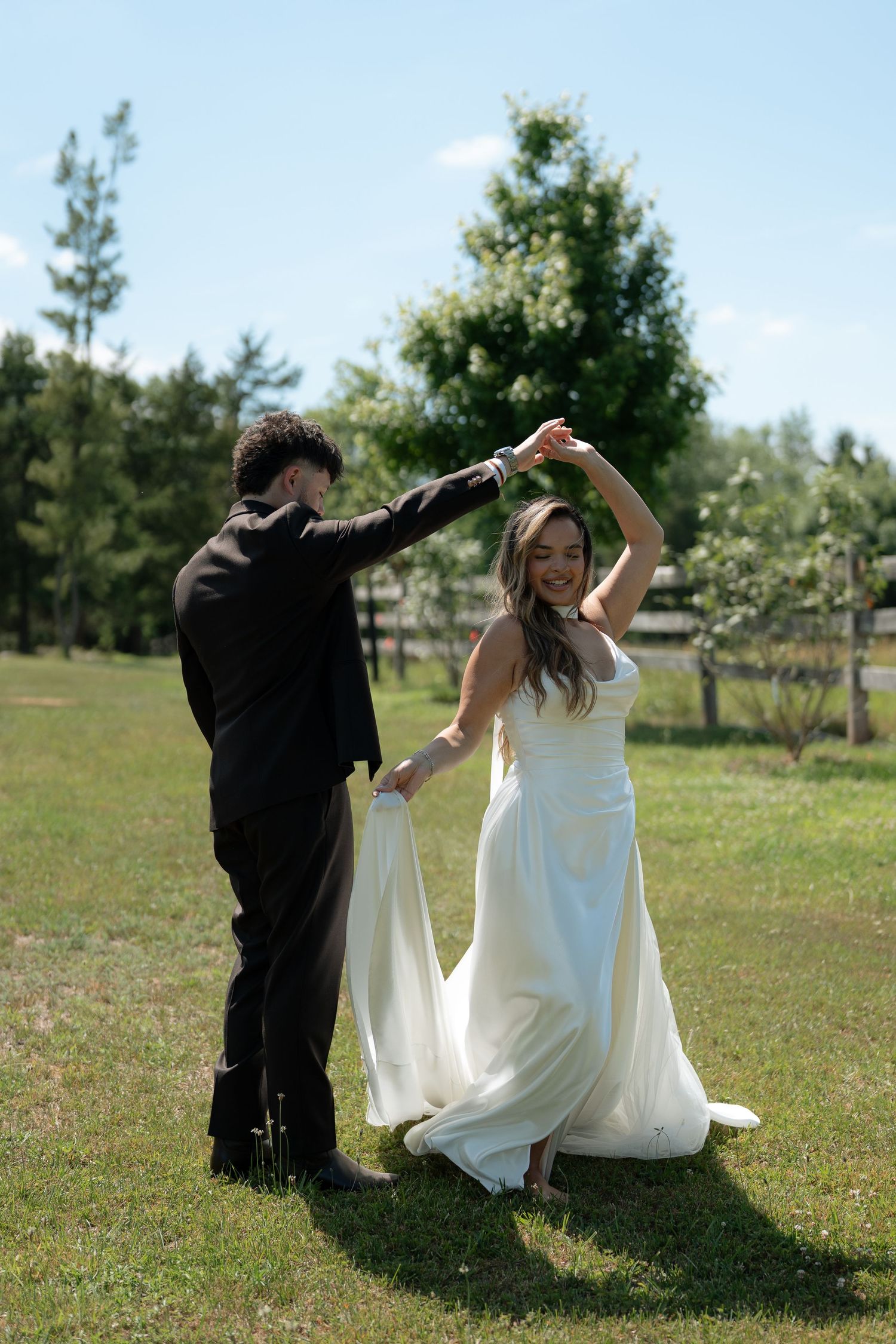 Bride and groom dancing outdoors on green grass, smiling joyfully in wedding attire on a sunny day.
