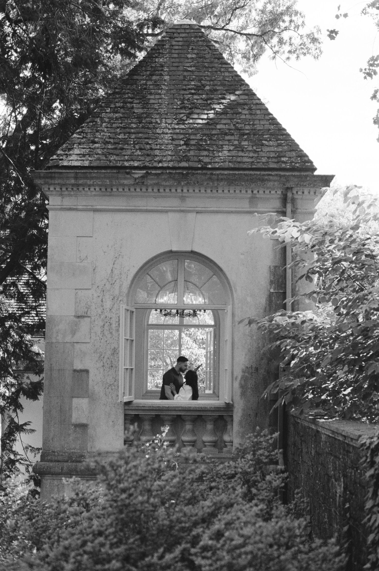 Black and white photo of a couple sharing a romantic kiss in an arched window of an elegant stone building.