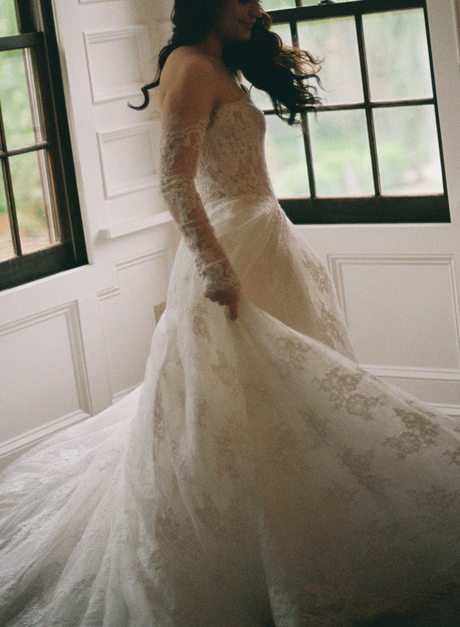 Bride in elegant lace wedding gown with long sleeves and flowing train standing by large windows.