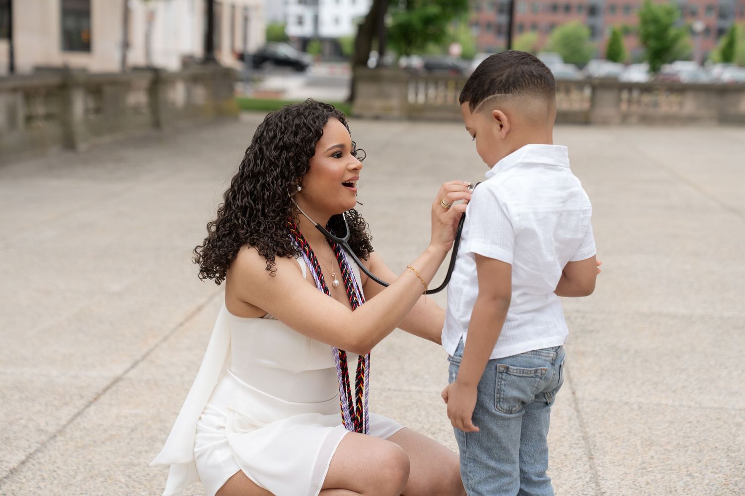 Young woman in white dress kneeling to adjust a stethoscope on a young boy in white shirt and jeans outdoors.
