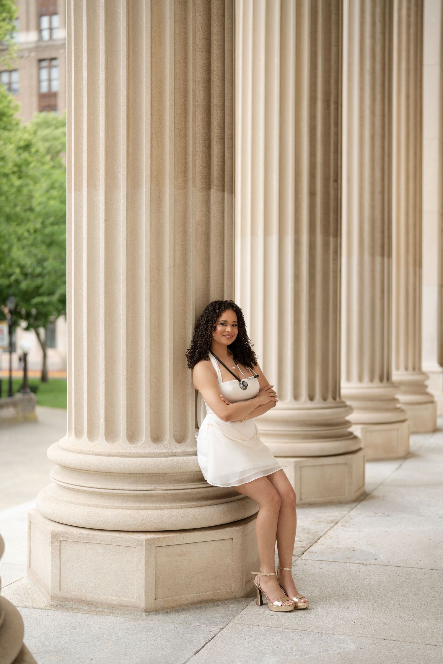 Young woman in white dress leans against classical stone columns smiling.
