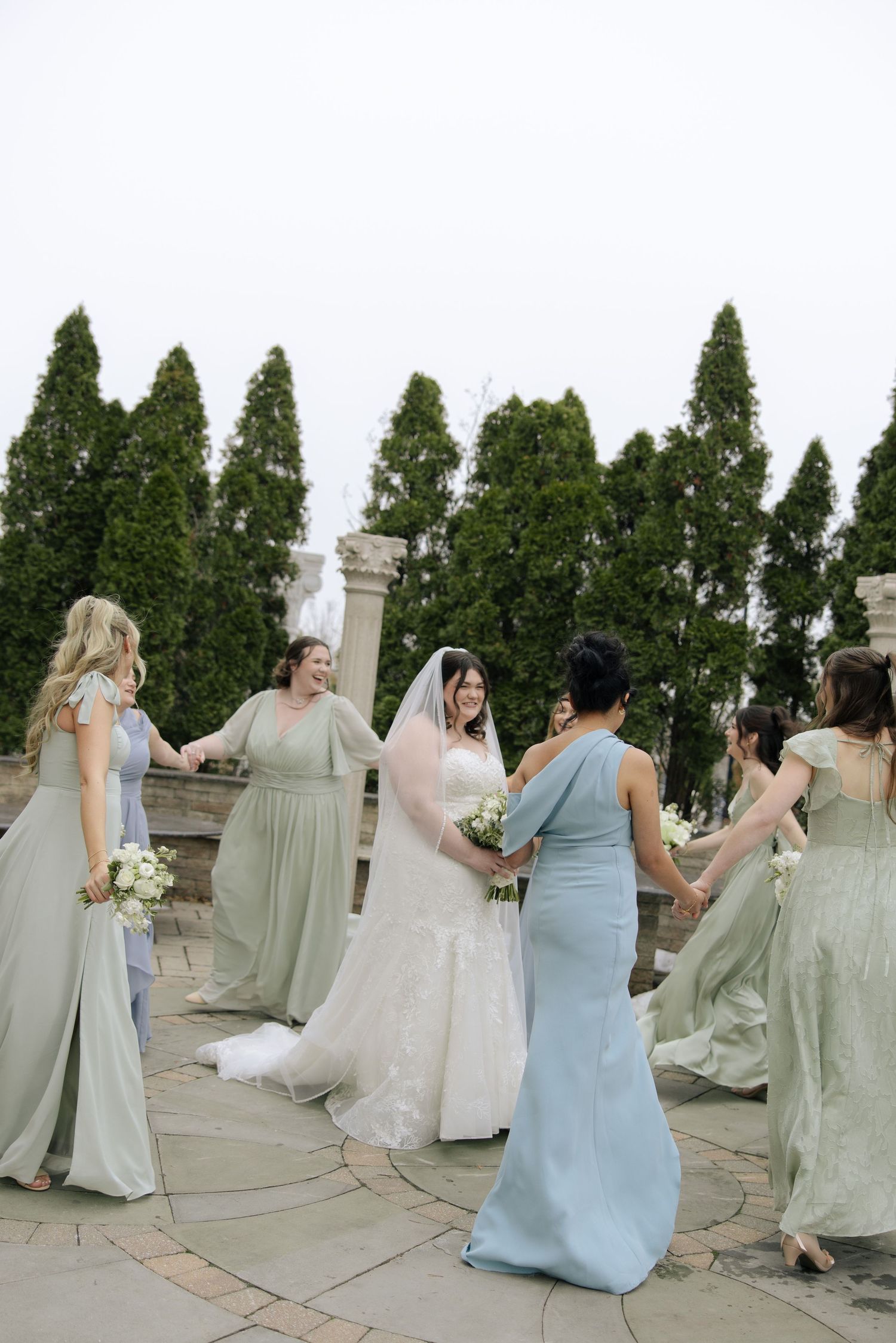 A bridal party in light blue and sage green dresses hold up their gowns while walking down a stone pathway.