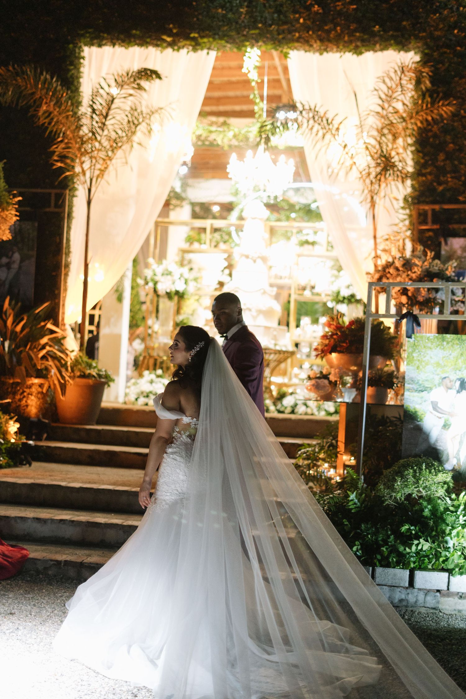A dramatic evening wedding photo with a long flowing veil and illuminated staircase in the background.