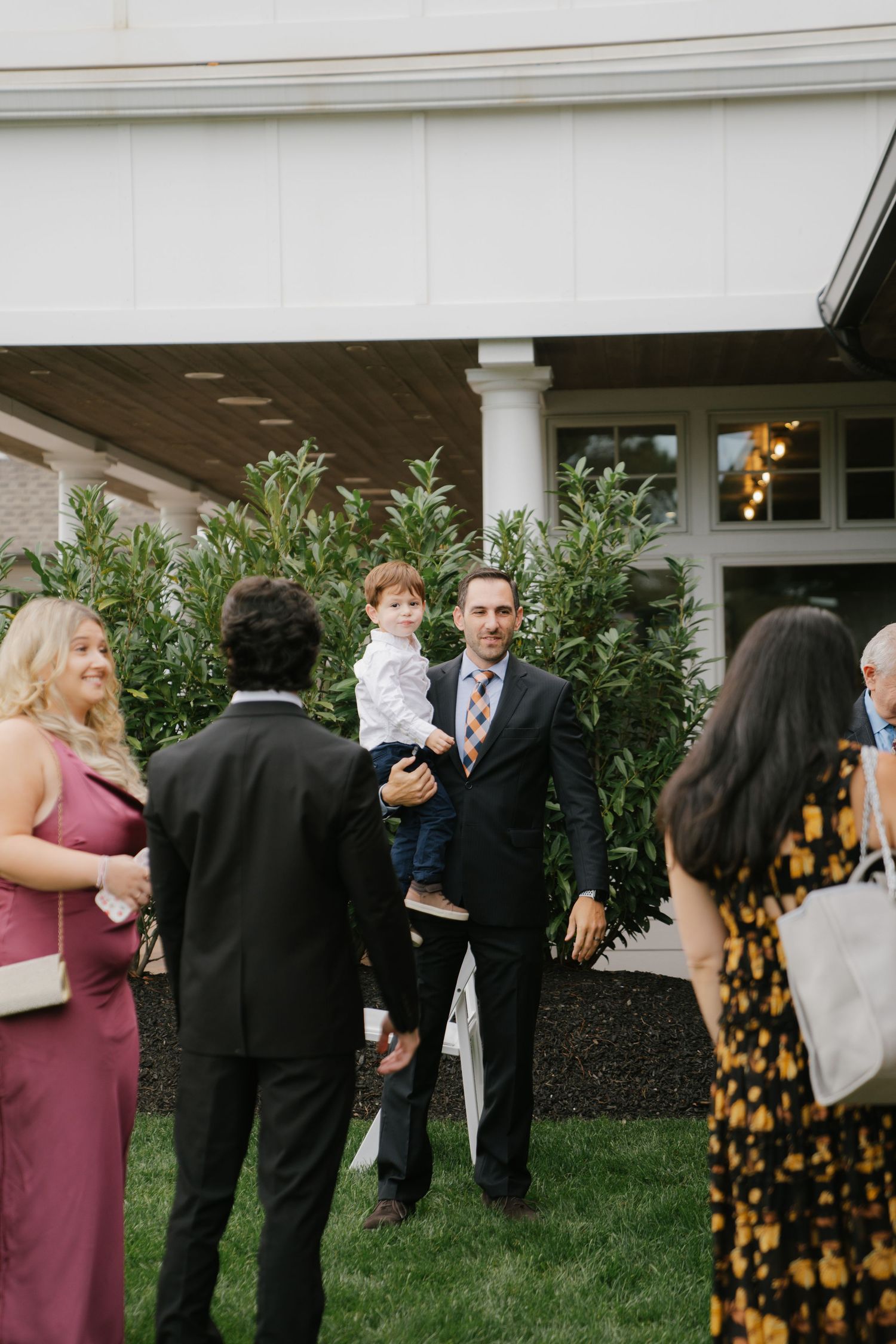 Wedding guests mingle and socialize on a manicured lawn outside a white-columned venue during an outdoor reception.