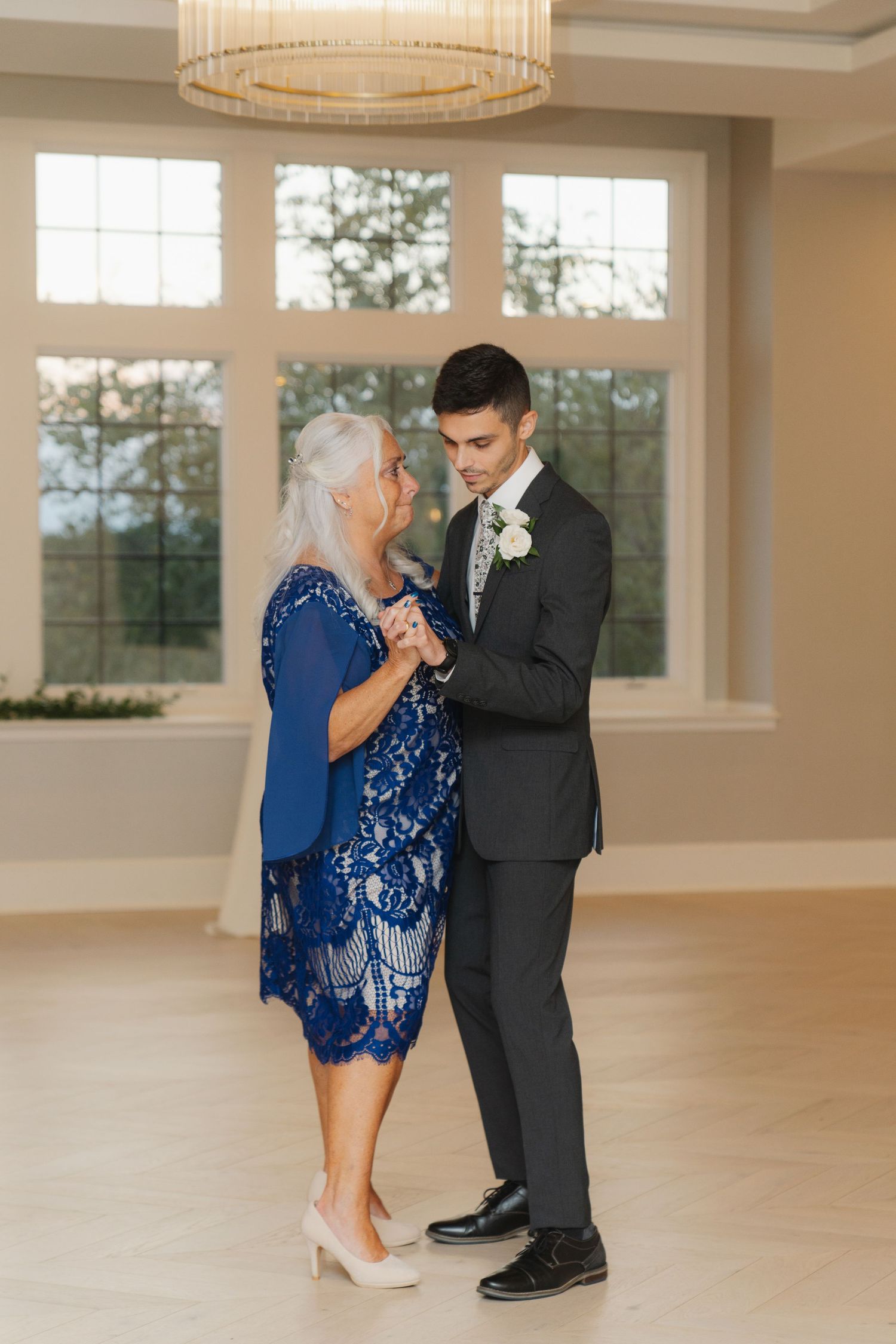 A touching dance moment between two people at a formal event in an elegant room with large windows and chandelier lighting.