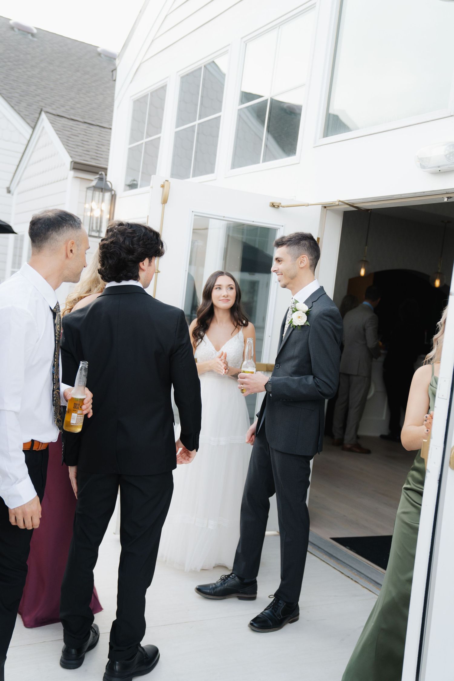 Wedding guests mingle and socialize on an outdoor patio with white walls and glass windows during a reception.