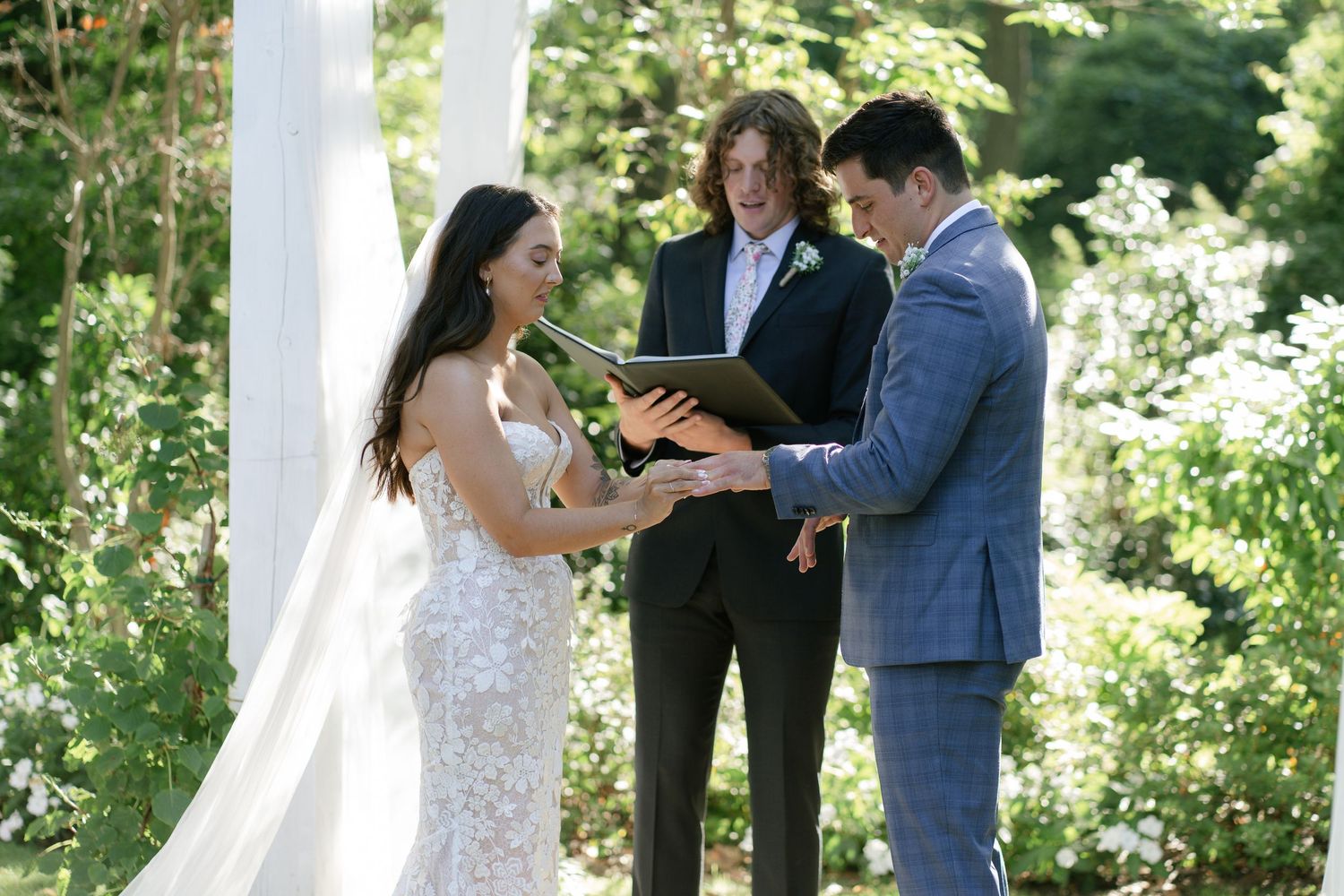 Wedding ceremony takes place under white pillars with lush greenery in the background.