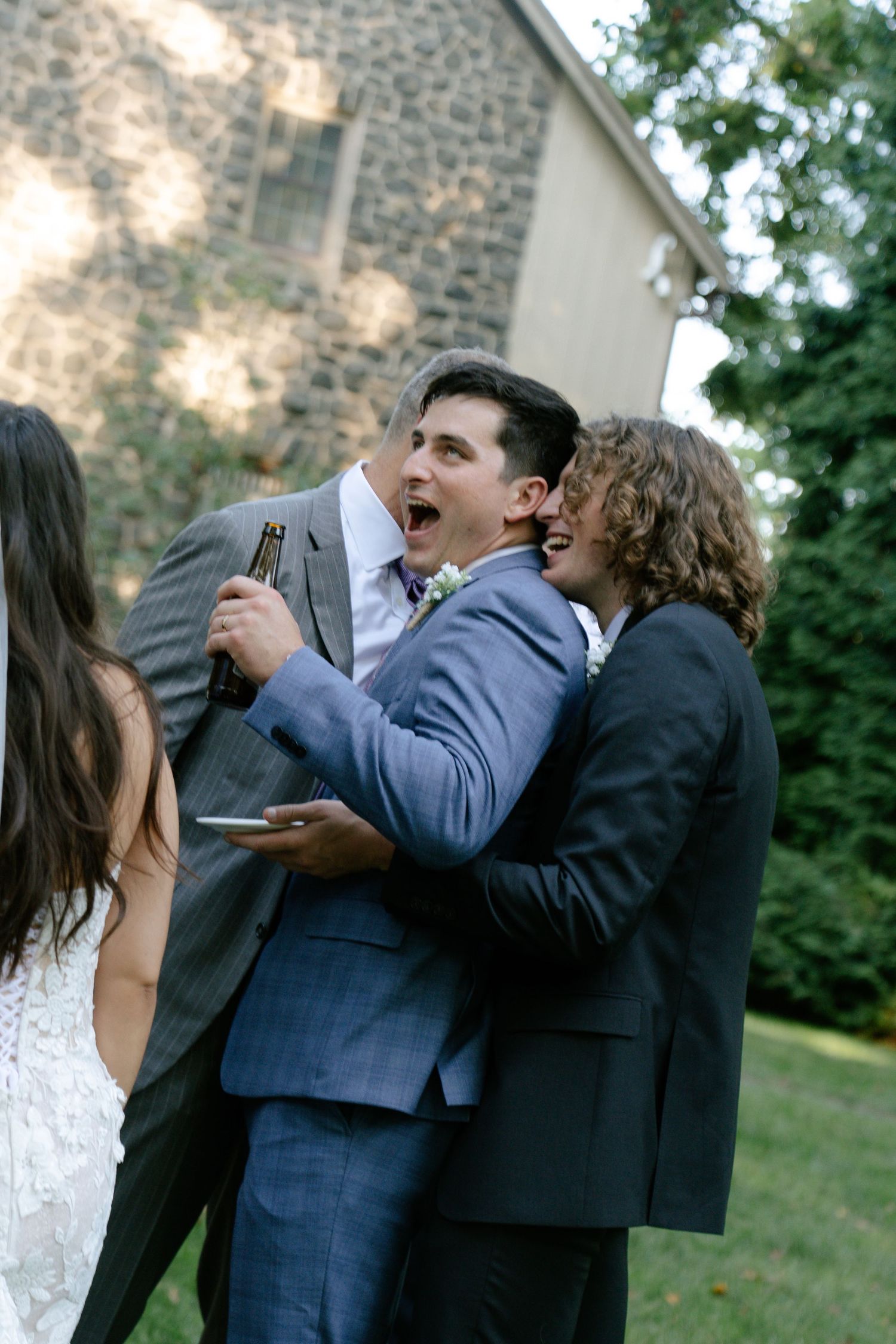 Wedding party members sharing laughs and embraces outside stone building.