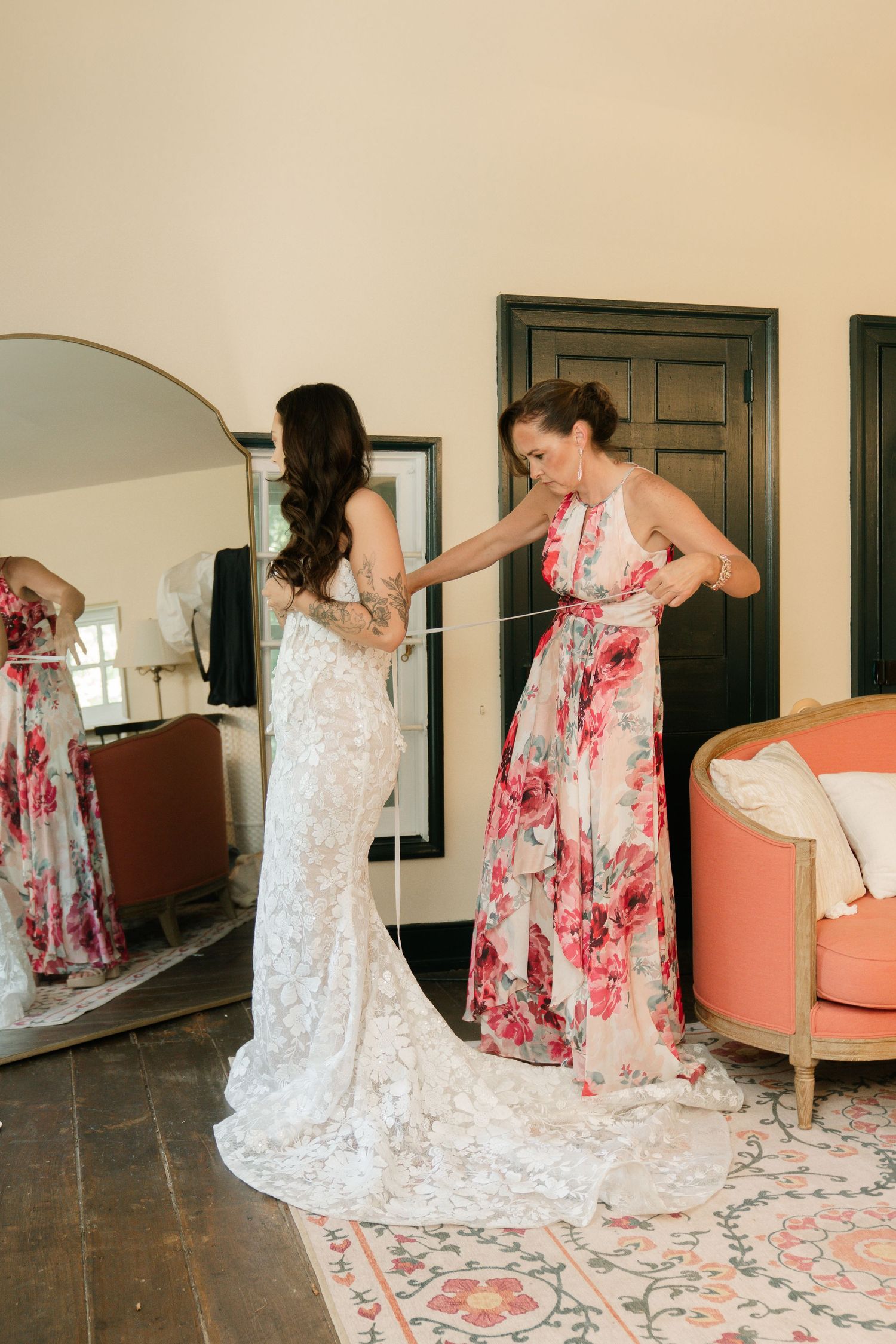Two women in elegant dresses, one white lace and one floral, dancing together in a sunlit room.