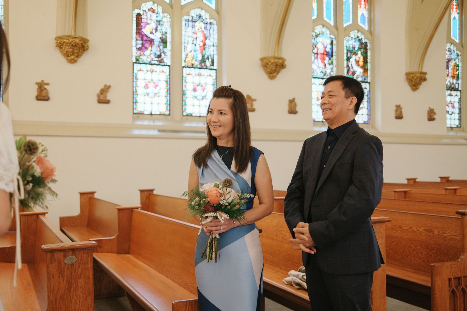 A couple stands together in a church with stained glass windows, wooden pews, and floral decorations.