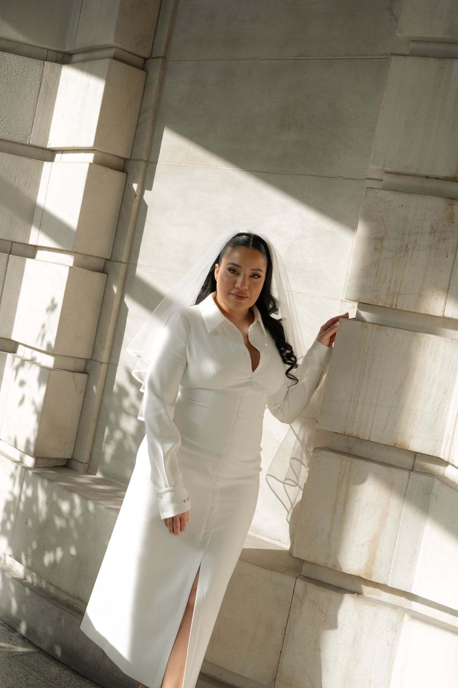 A person in a white dress poses against a wall with dramatic shadows and lighting.