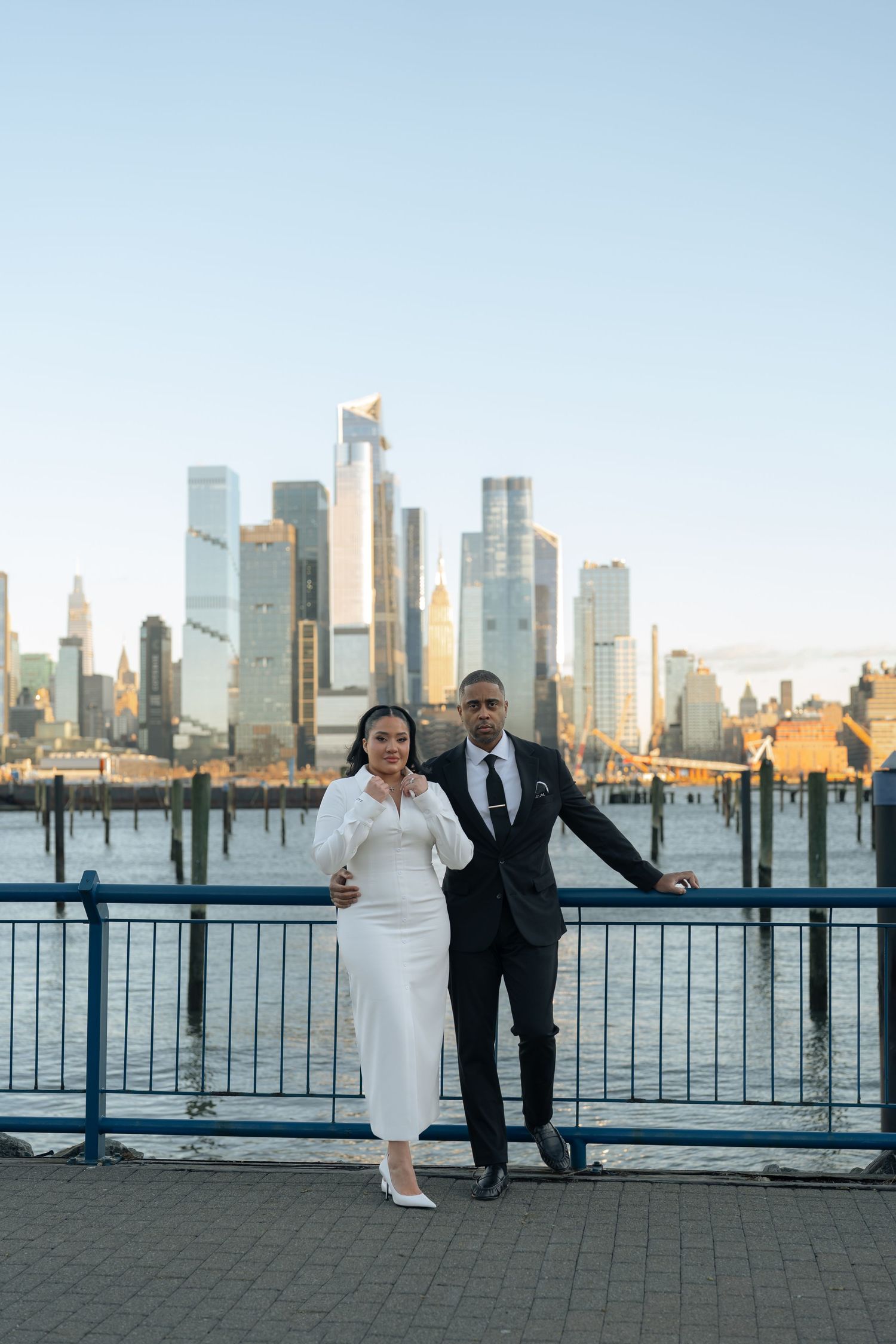 Newlyweds pose playfully against a railing with Manhattan's skyline creating a stunning backdrop.