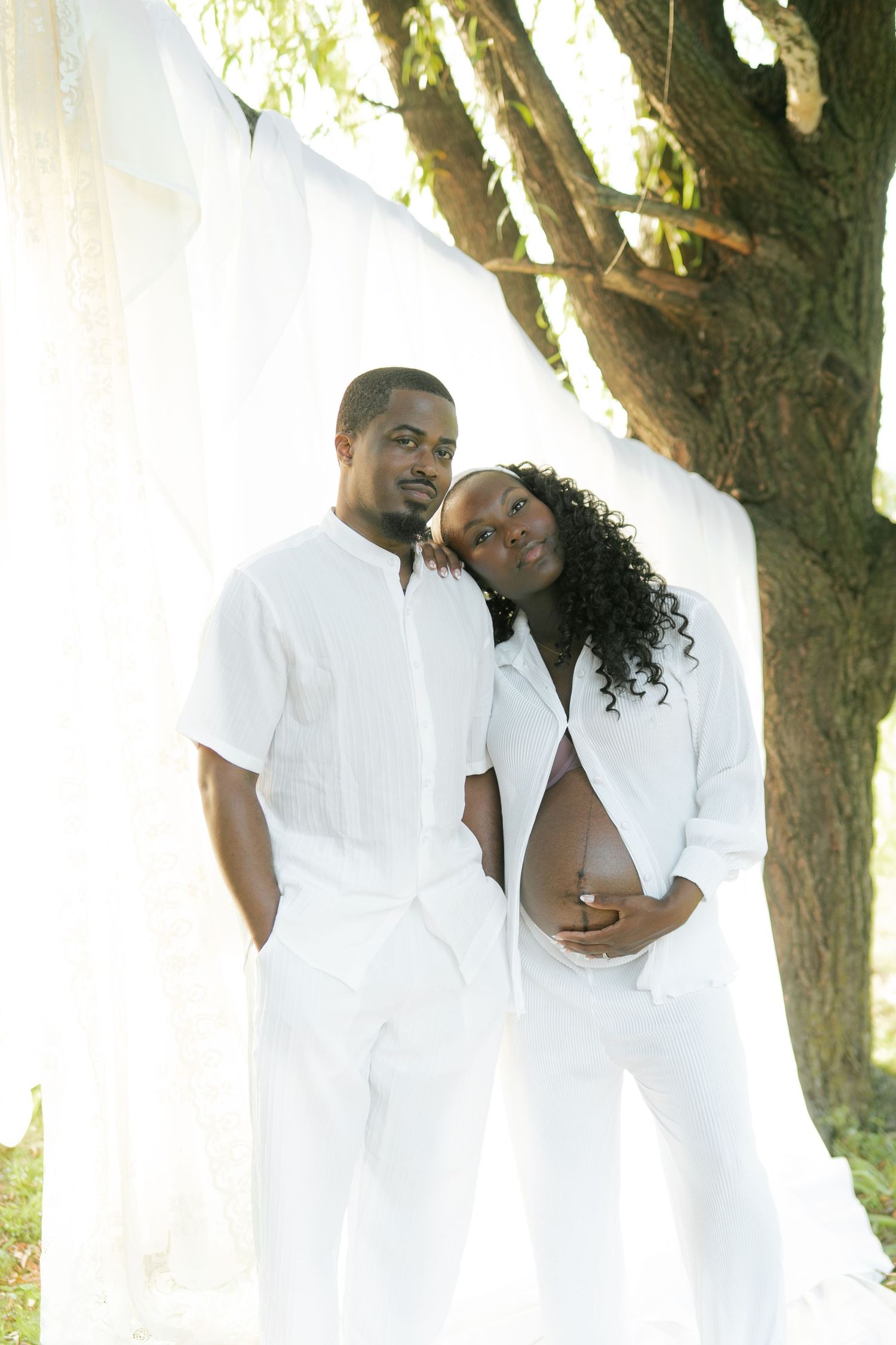 A pregnant couple dressed in white poses outdoors under a tree with flowing white fabric backdrop.