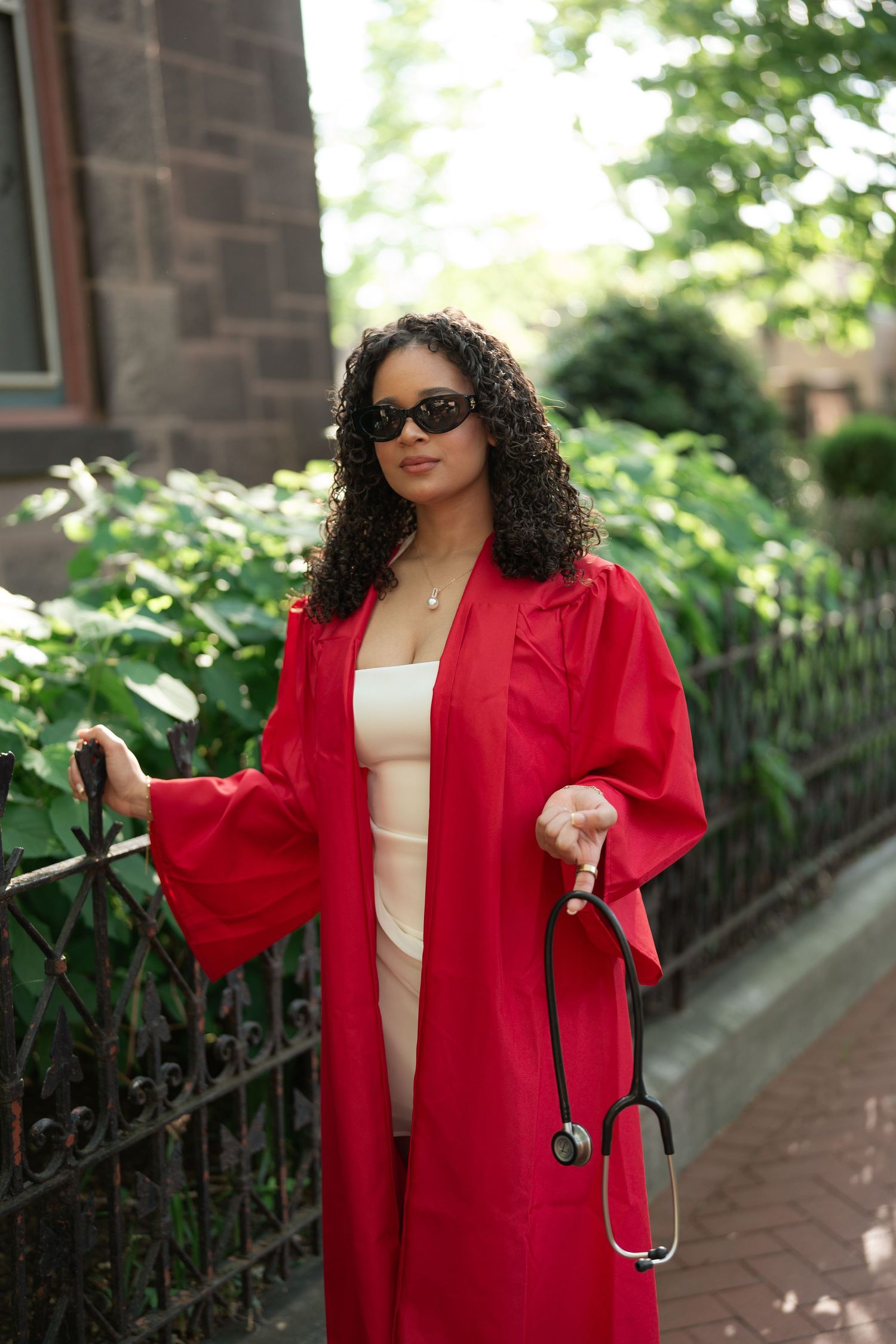 Graduate in red cap and gown holds stethoscope outdoors, wearing sunglasses near iron fence.