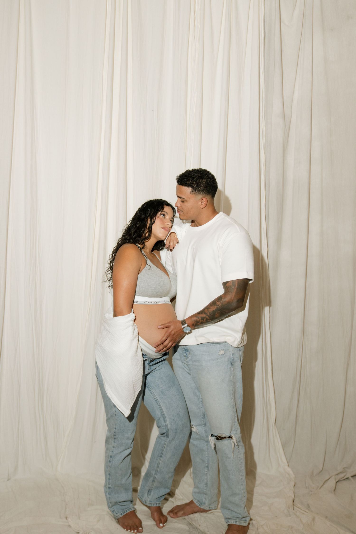Pregnant couple in jeans posing together against white curtain backdrop, woman in crop top showing baby bump.