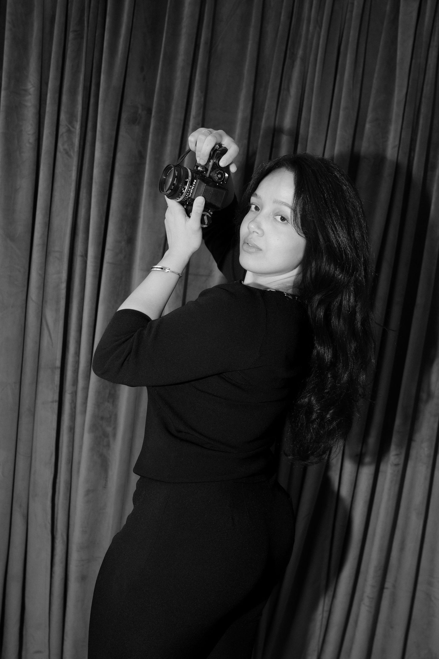 Black and white photo of a young woman in black outfit holding a vintage camera against draped curtain backdrop.