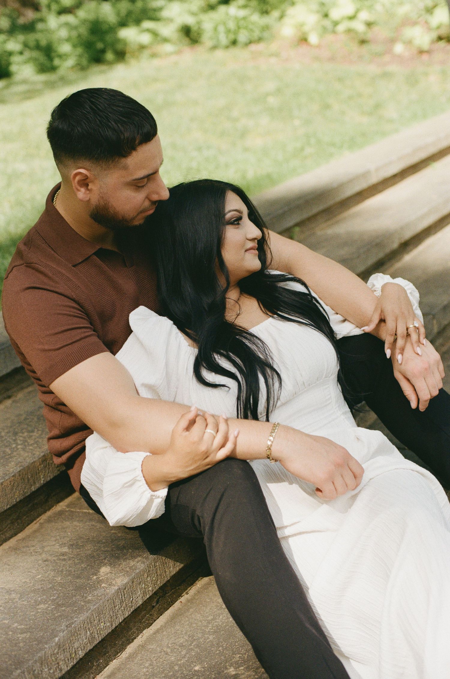 Couple sitting on stone steps in a park, woman in white dress leaning against man in brown shirt.
