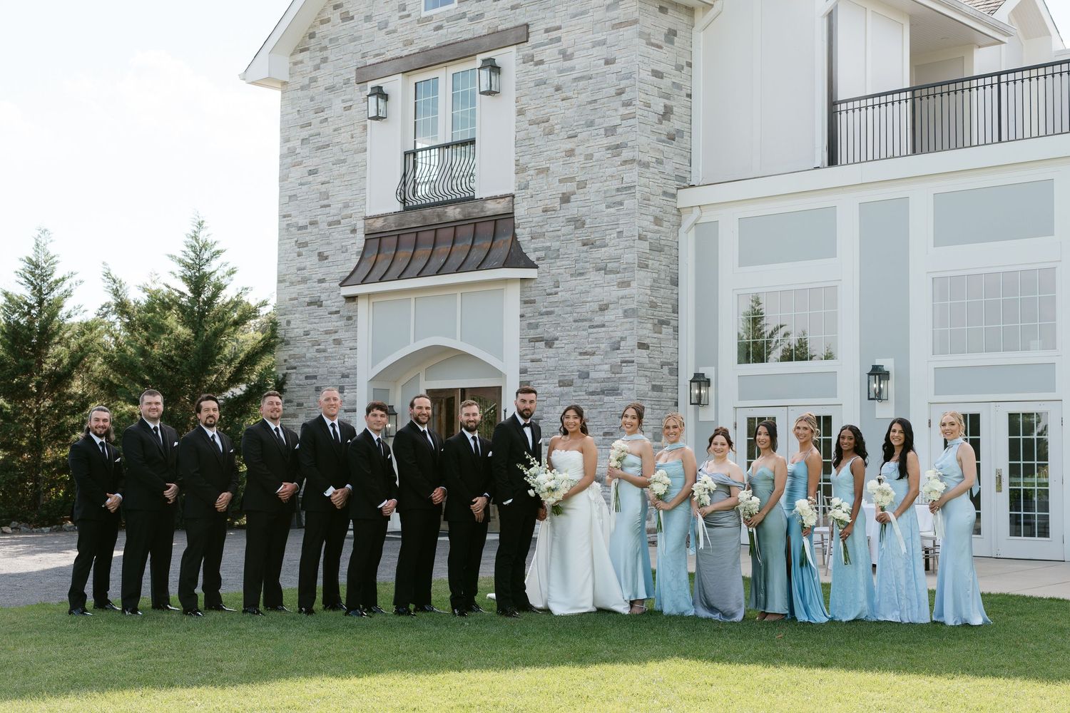 Large wedding party poses outside stone venue, groomsmen in black suits and bridesmaids in light blue gowns.