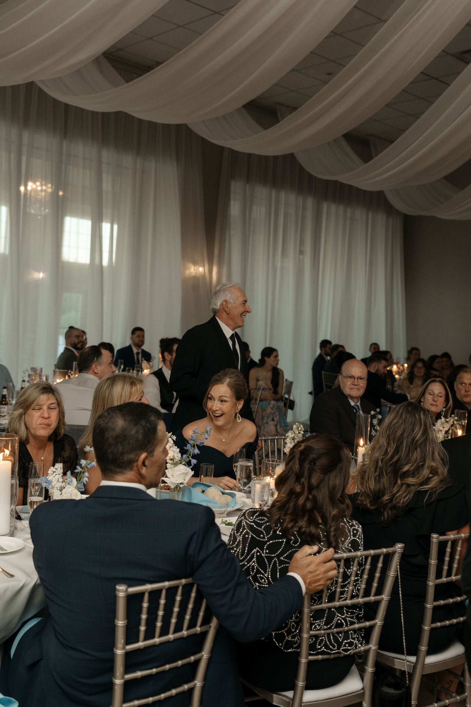 Wedding guests laugh and celebrate during elegant reception dinner under draped white ceiling.
