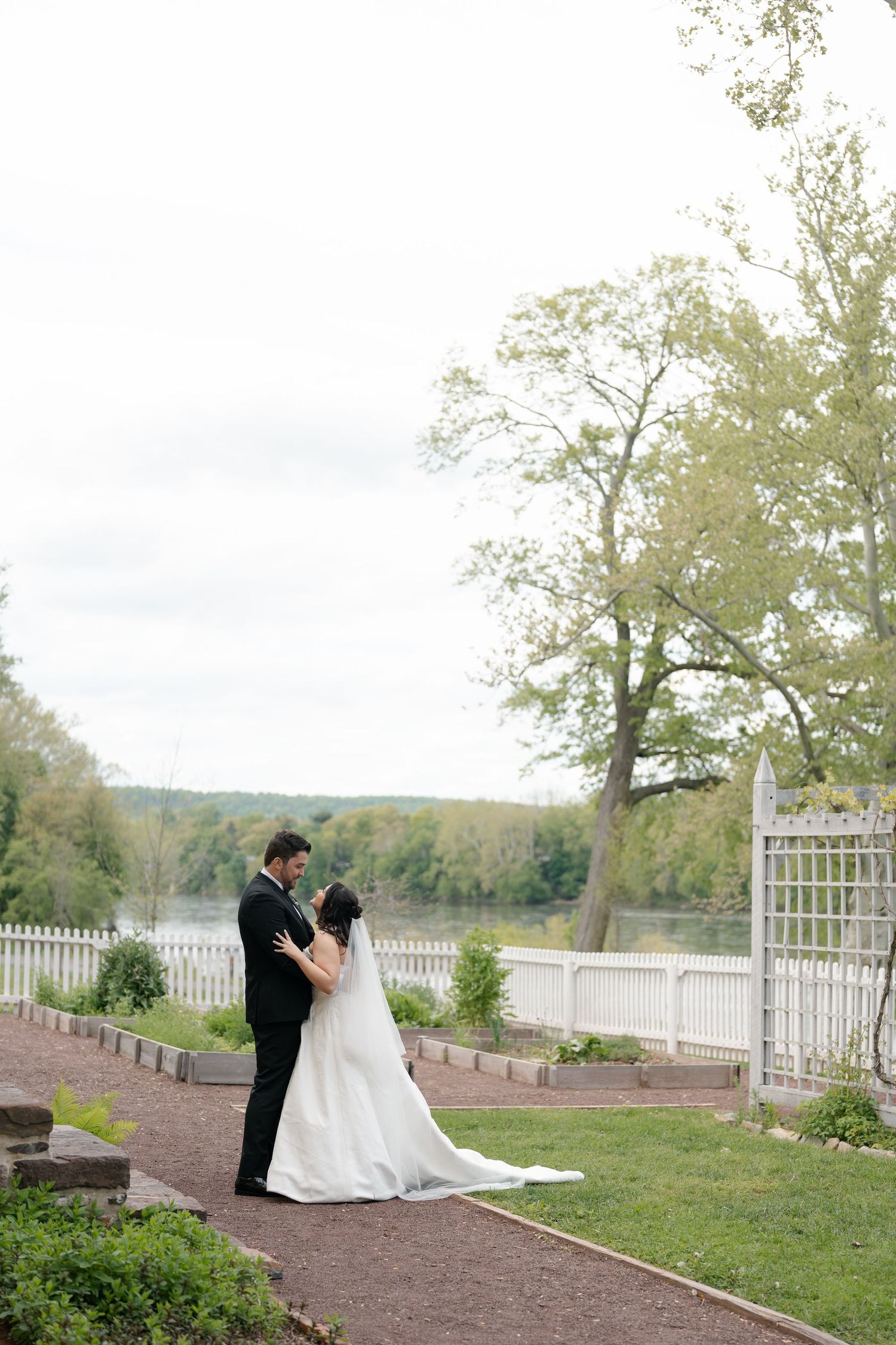Bride and groom share a romantic embrace in a garden by a serene river, surrounded by lush trees and a white fence.
