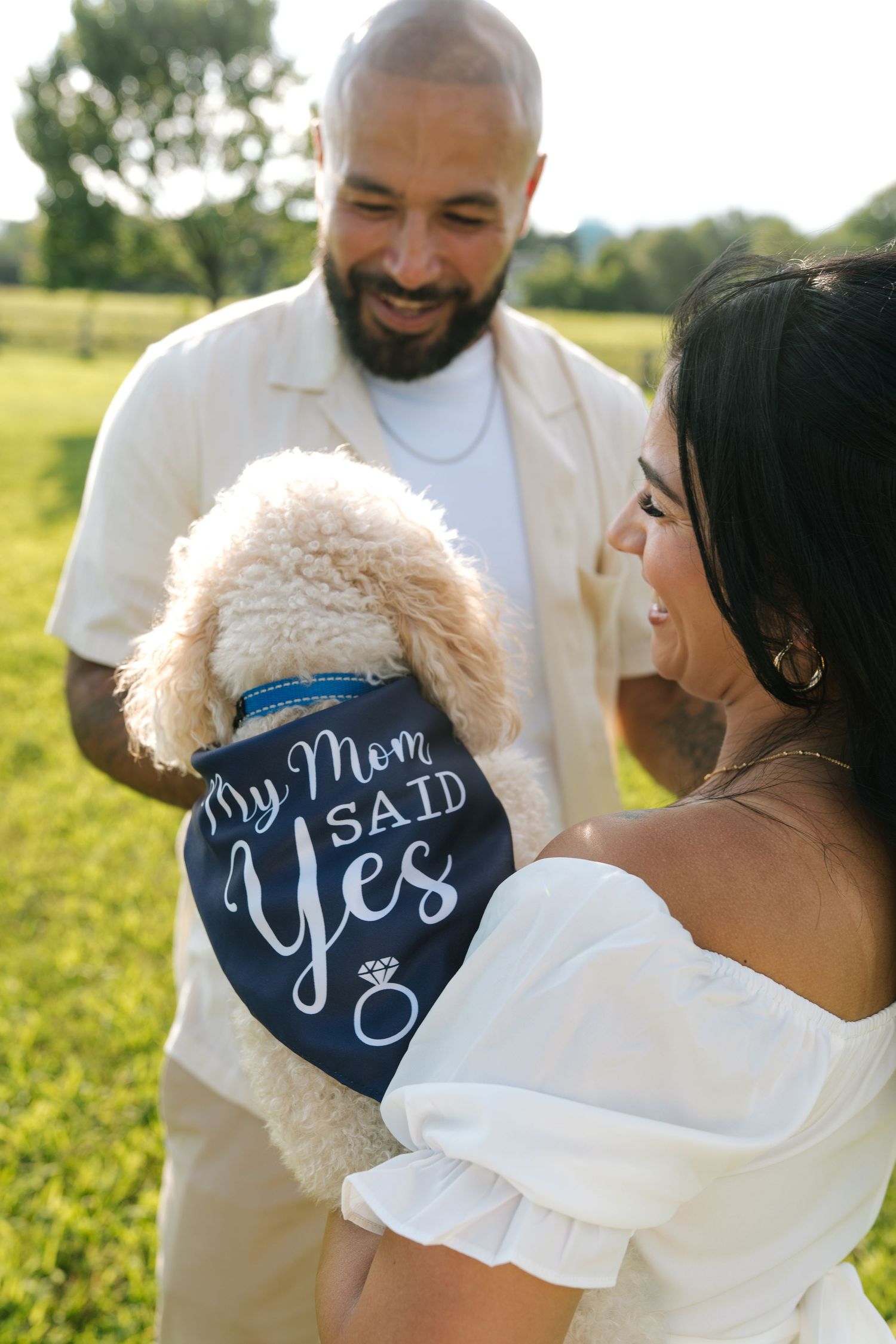 A dog wearing a navy blue bandana with white text sits between a couple during an outdoor photoshoot.