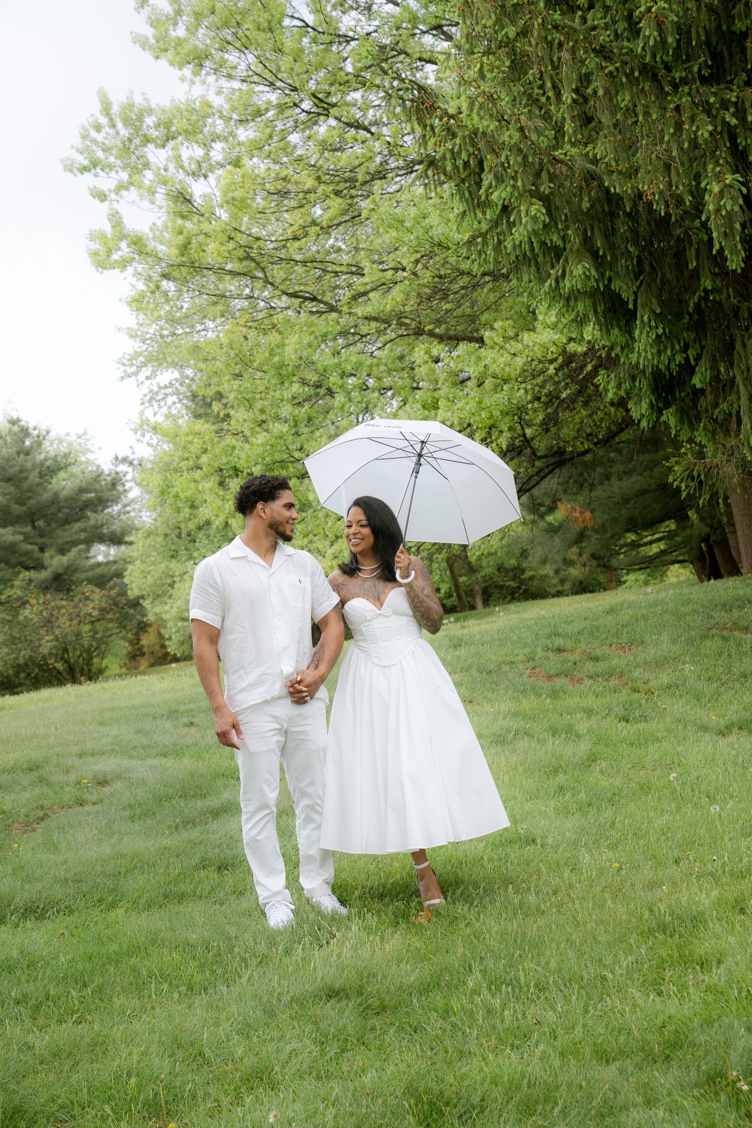 Couple in white outfits sharing a romantic moment under an umbrella in a lush green park.