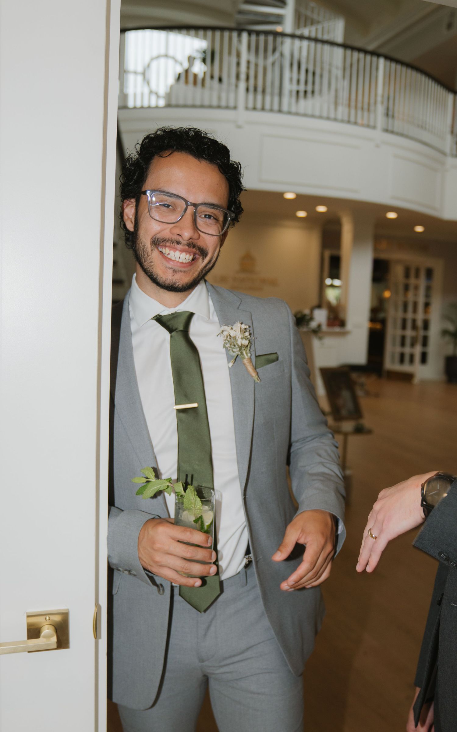 A wedding guest in a grey suit and green tie smiles while holding a flower at an elegant indoor venue.