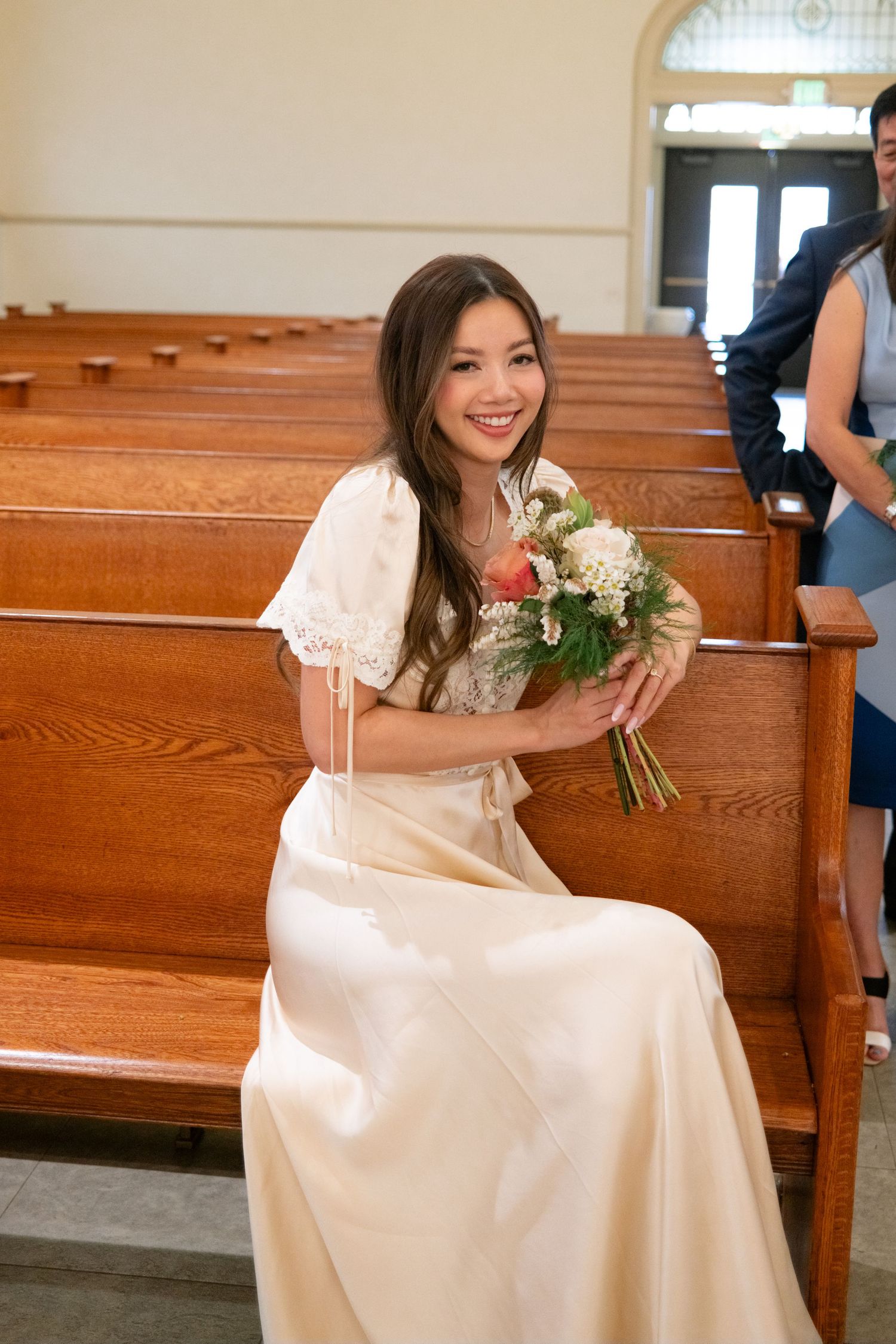 A bride in a cream-colored dress sits on a wooden church pew holding a bouquet of white and pink flowers.