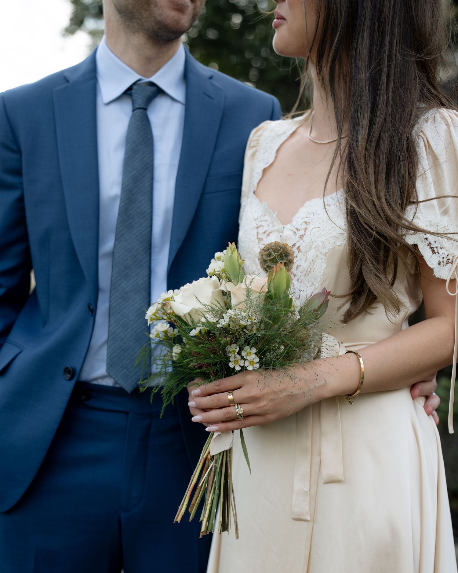 Close-up details of blue suit and cream dress with floral bouquet during wedding ceremony.