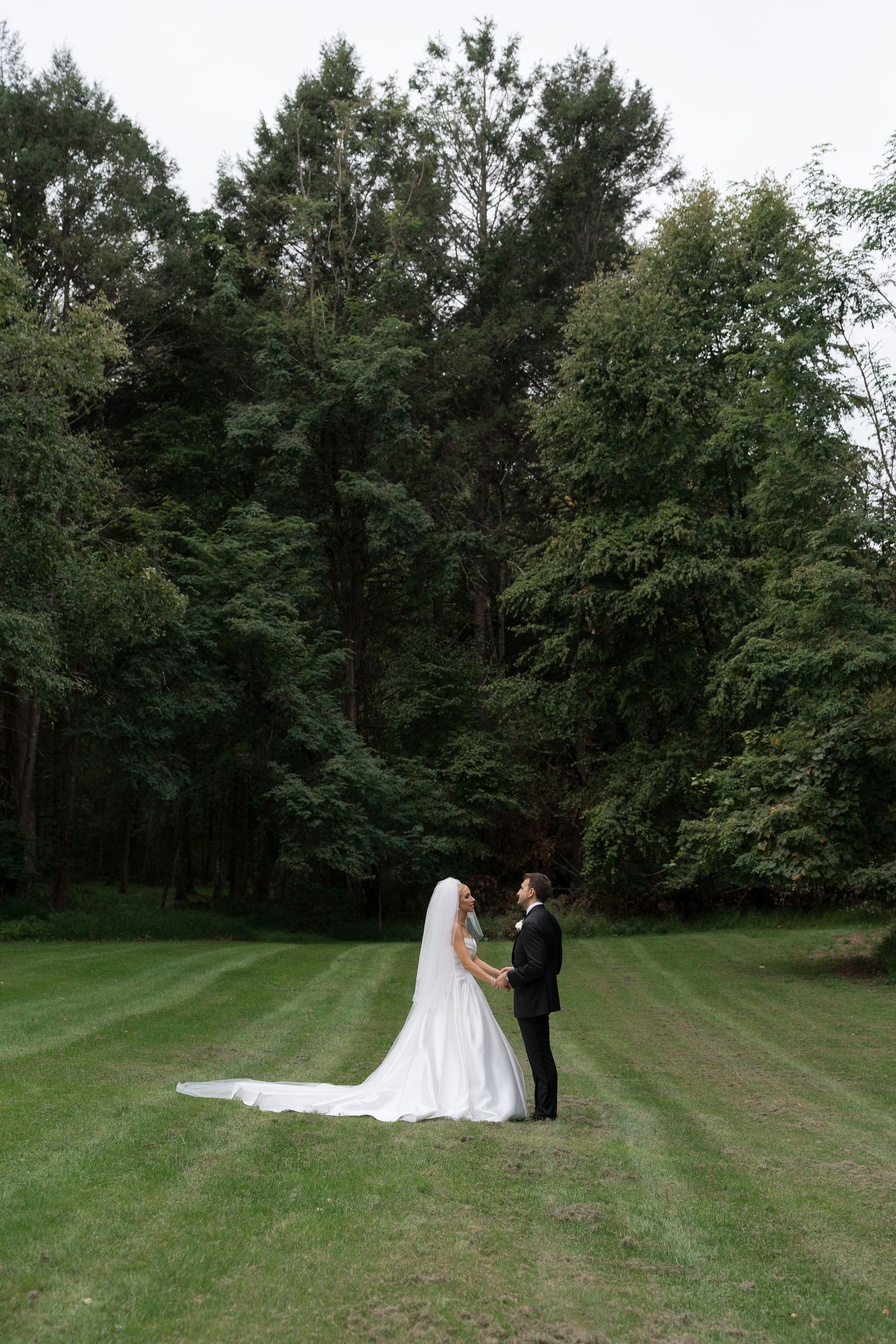 Bride and groom share a romantic moment on a lush green lawn surrounded by tall trees.