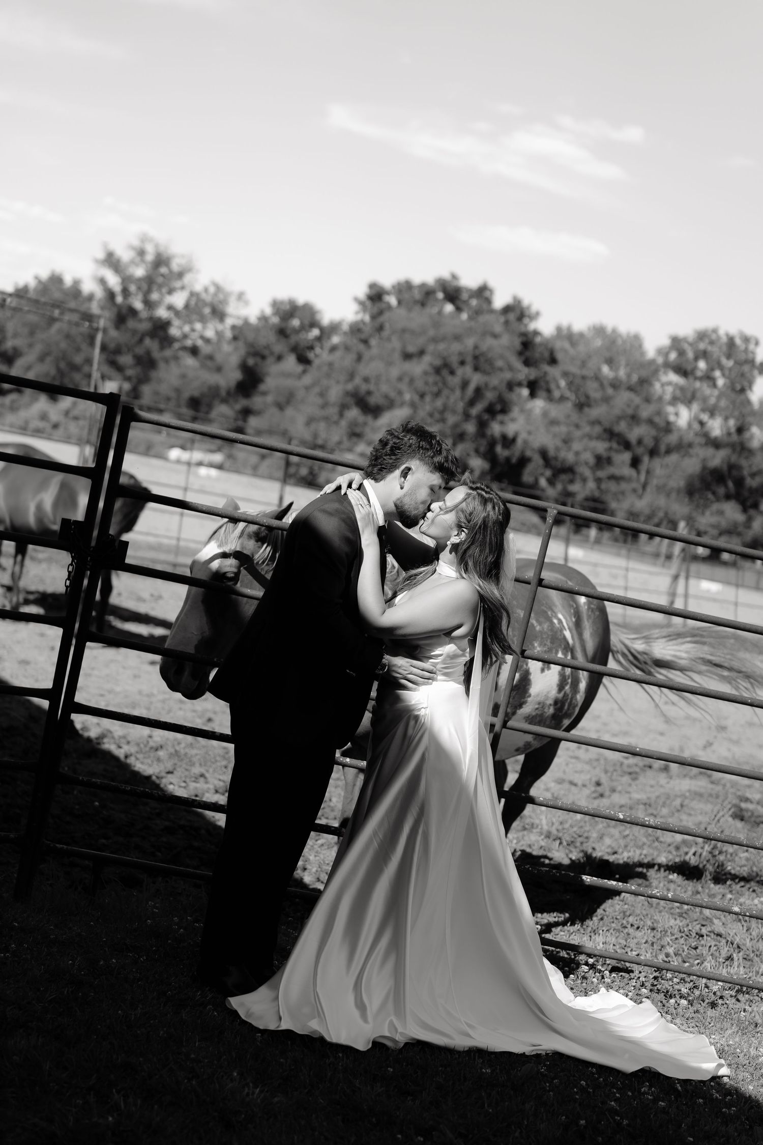 Black and white photo of bride and groom sharing romantic moment near farm fence with horses.