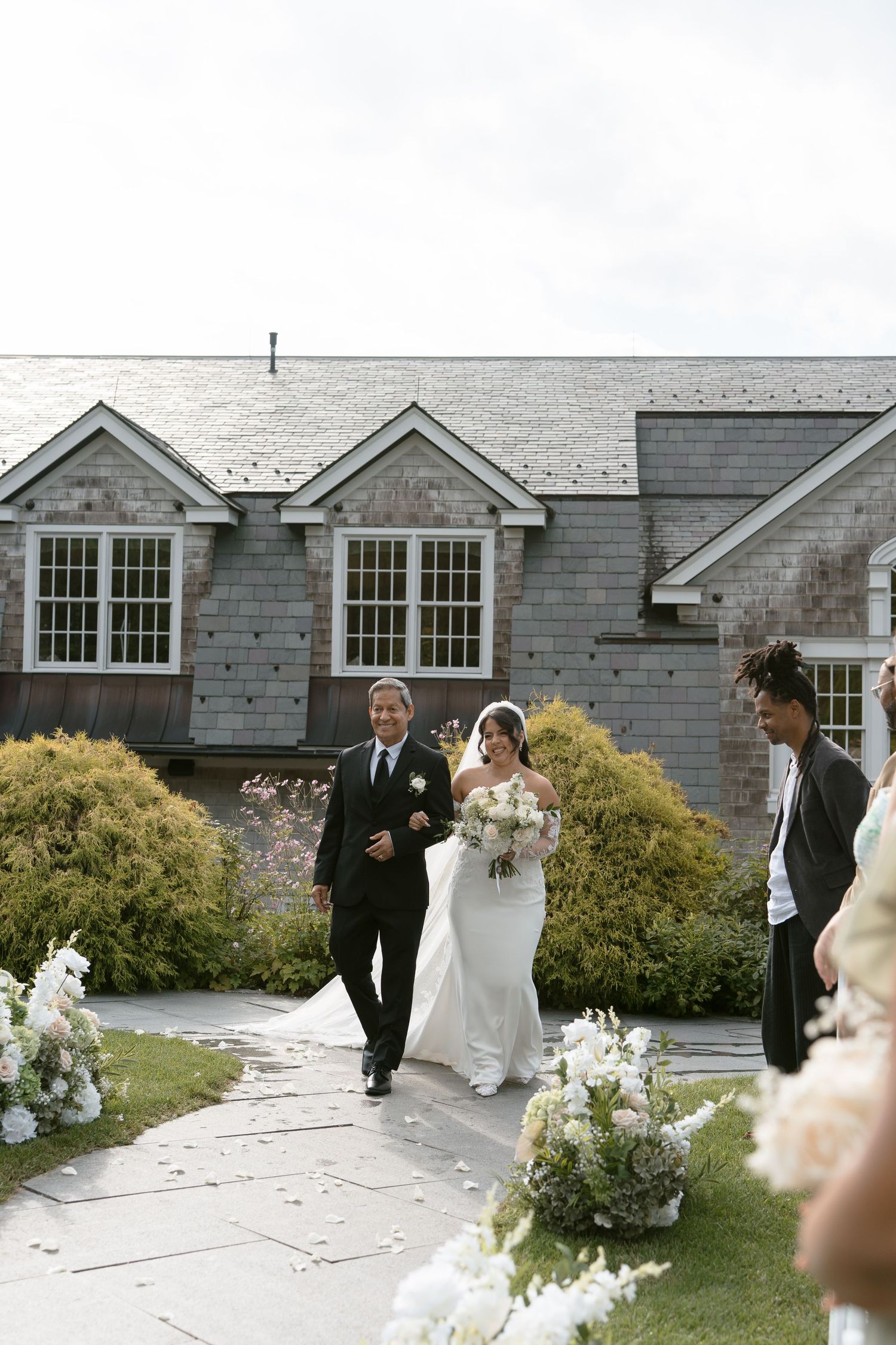 Bride walks down outdoor aisle with escort past white floral arrangements at elegant wedding ceremony.