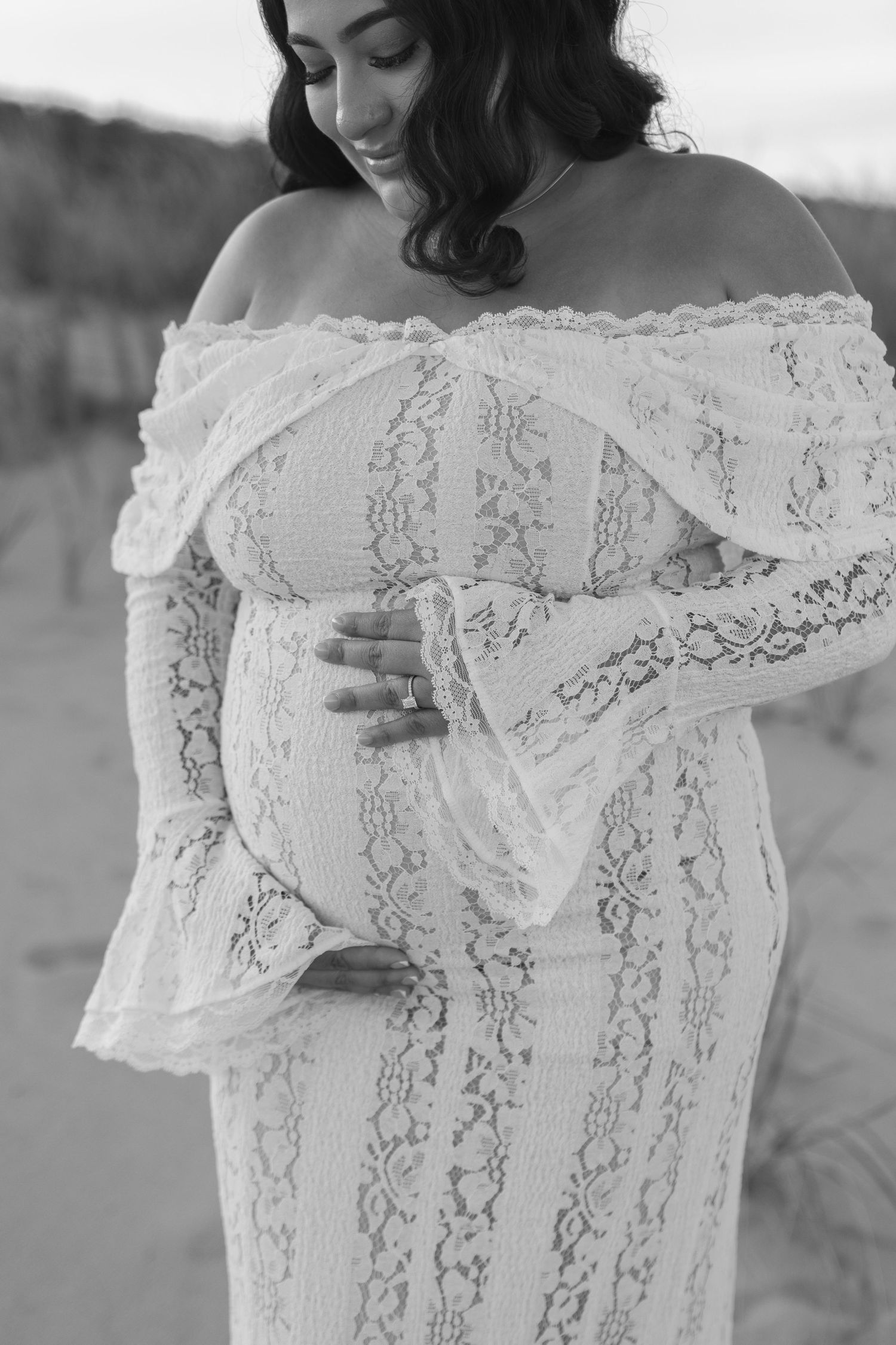 Black and white maternity photo of a smiling woman in an off-shoulder lace gown cradling her baby bump.