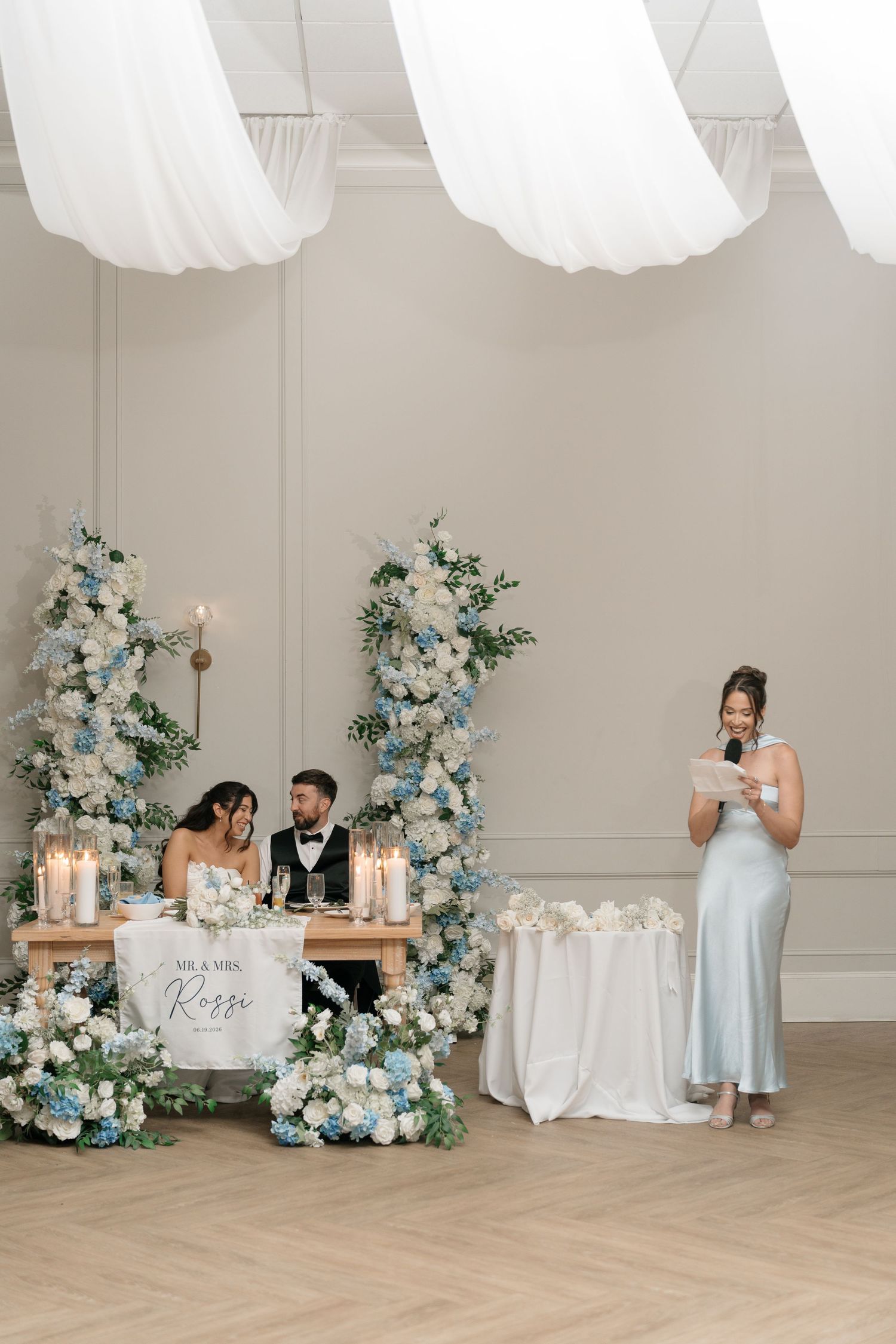 Elegant wedding reception sweetheart table with blue and white floral arrangements while bridesmaid delivers speech.