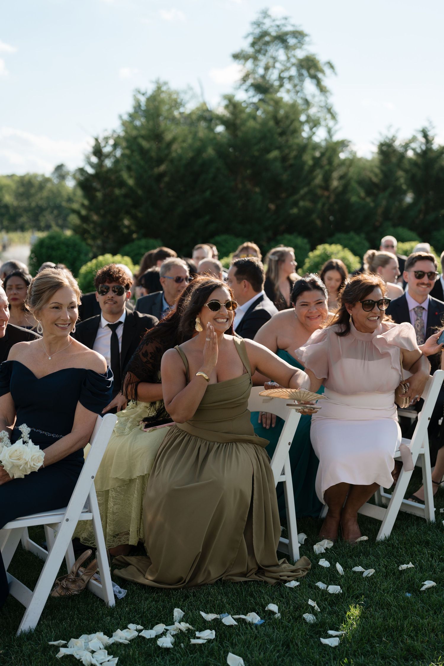 Smiling wedding guests seated in white chairs at an outdoor ceremony on a sunny day.
