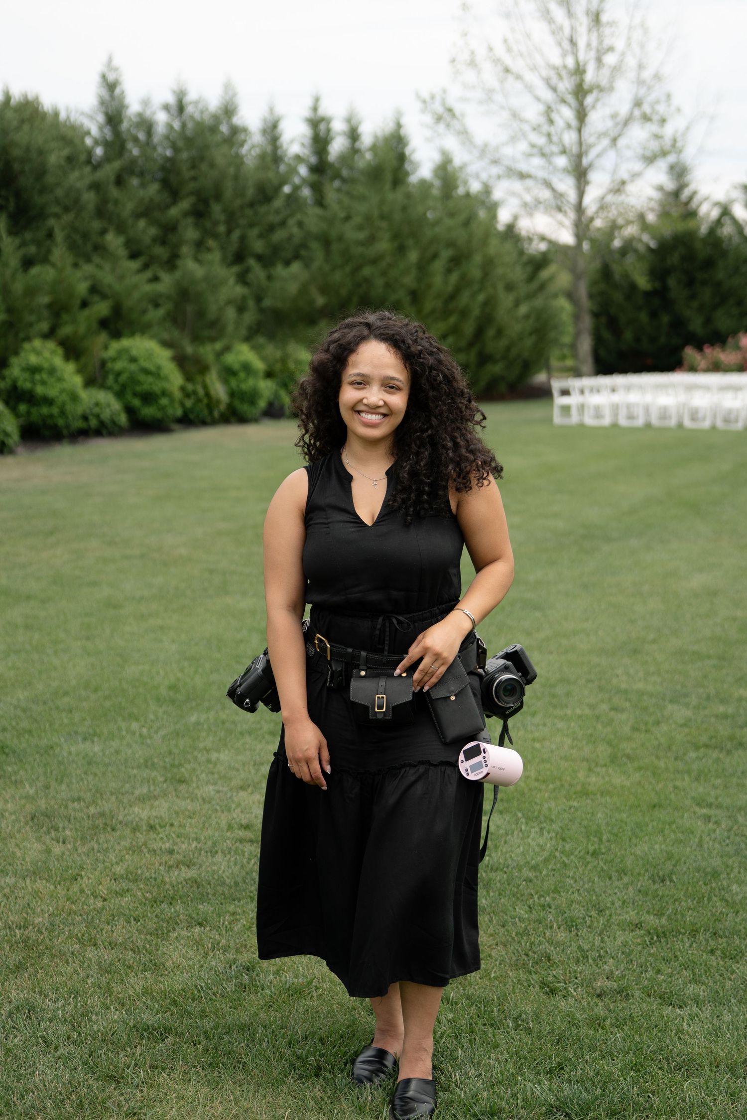 A smiling woman in a black dress holds cameras while standing on a green lawn at an outdoor event venue.