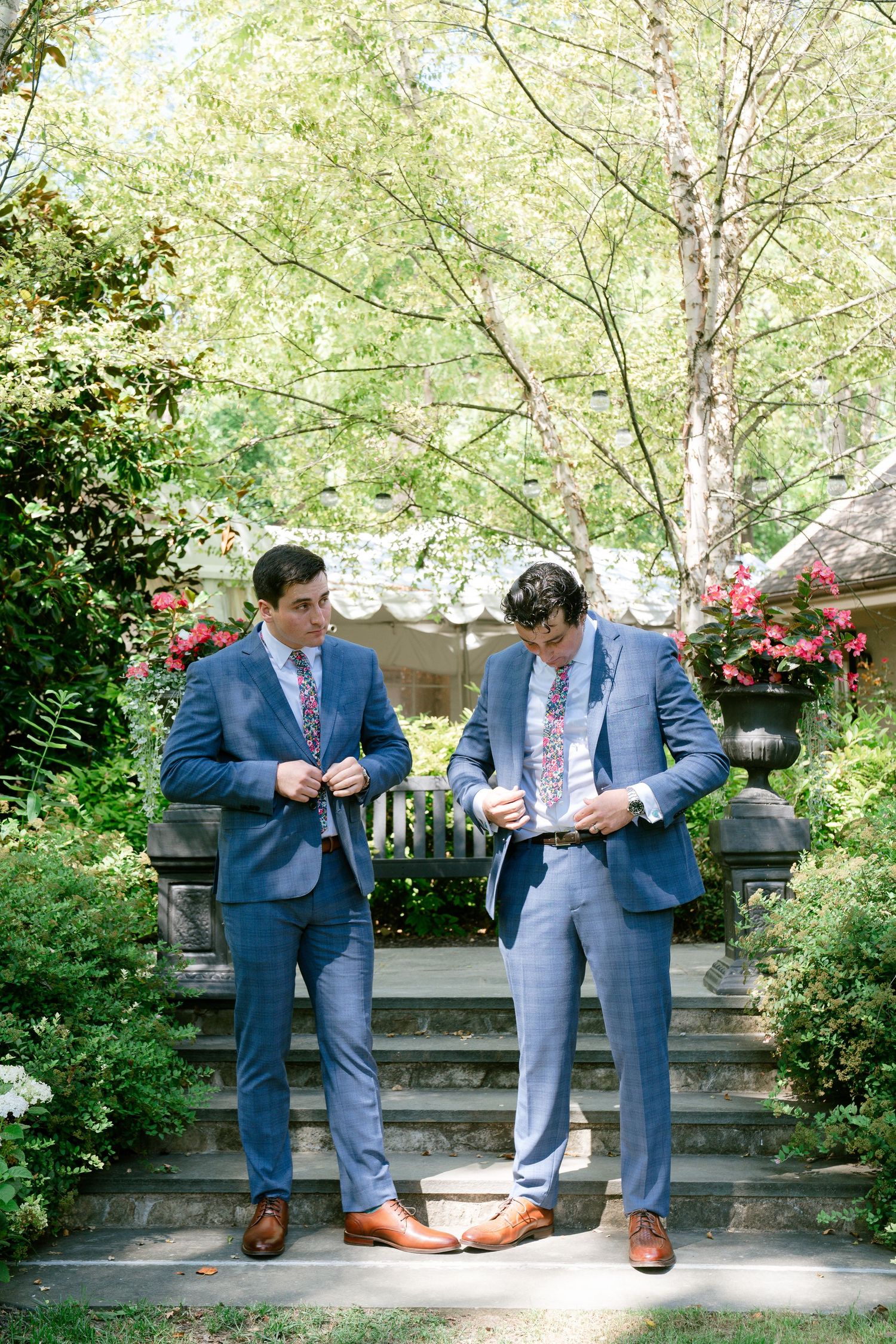 Two men in blue suits stand on stone garden steps surrounded by greenery and flowers.