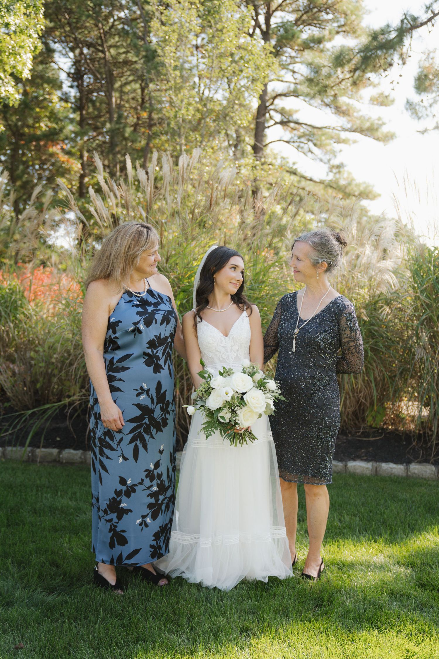 Three people stand together on grass near evergreen trees during an outdoor wedding celebration.