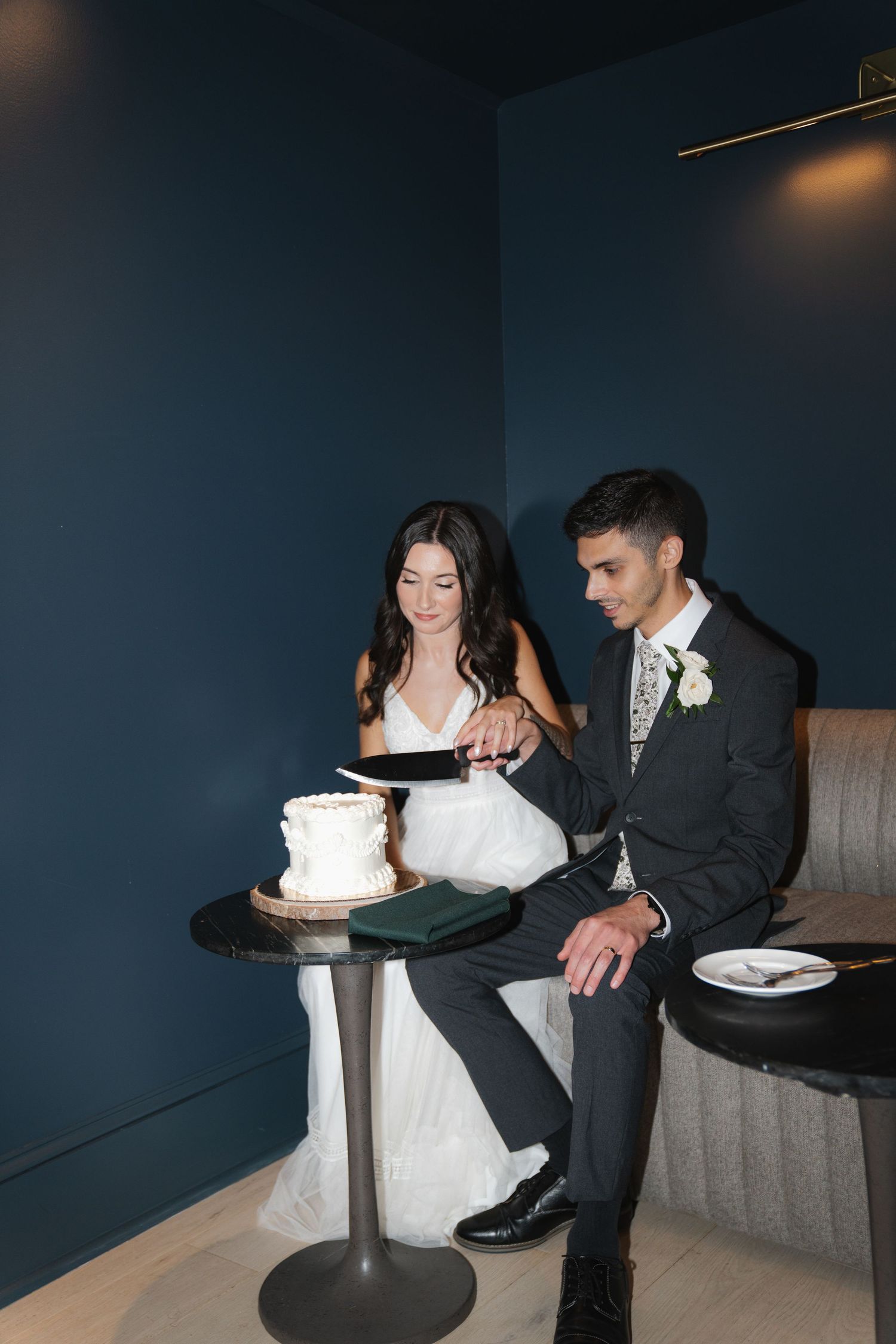 A bride and groom in formal wedding attire cut their white wedding cake together during their reception.