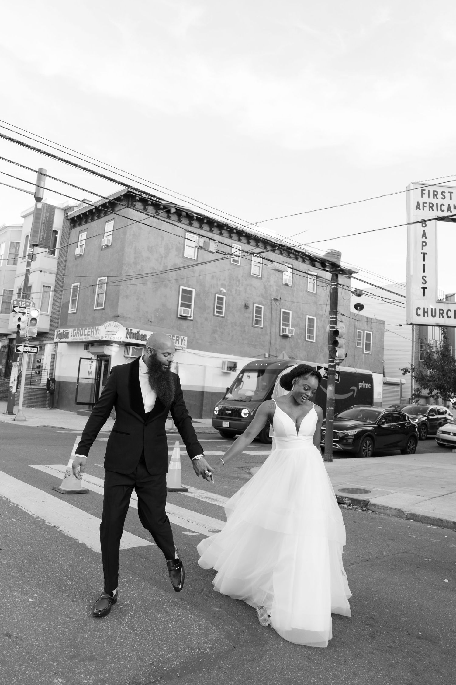 A couple in wedding attire walks together on a street with a church visible in the background.
