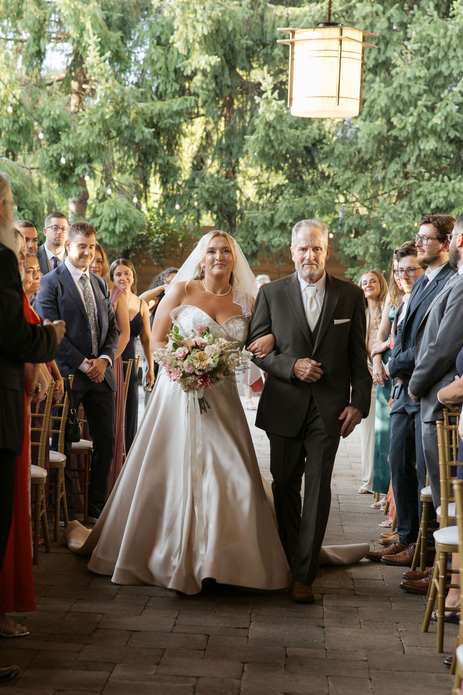 Bride walks down outdoor aisle with escort holding pink bouquet in satin ball gown with veil at wedding ceremony.