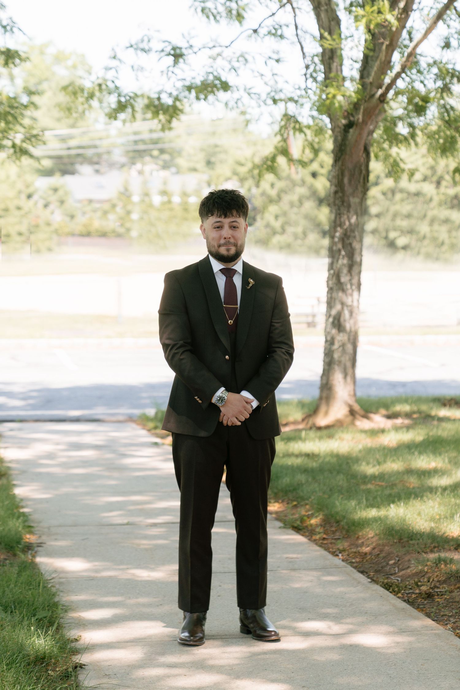 Groom in dark brown three-piece suit with burgundy tie standing on outdoor pathway at wedding.