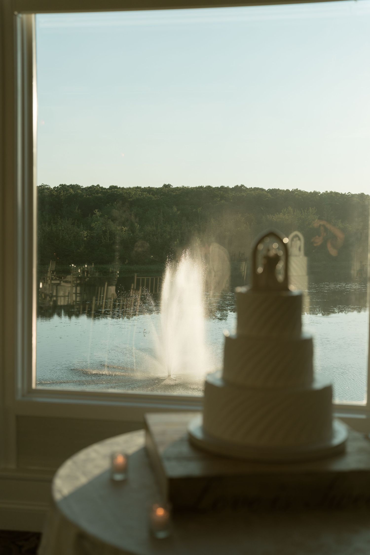 Three-tier white wedding cake displayed on a table with a scenic lake fountain view.