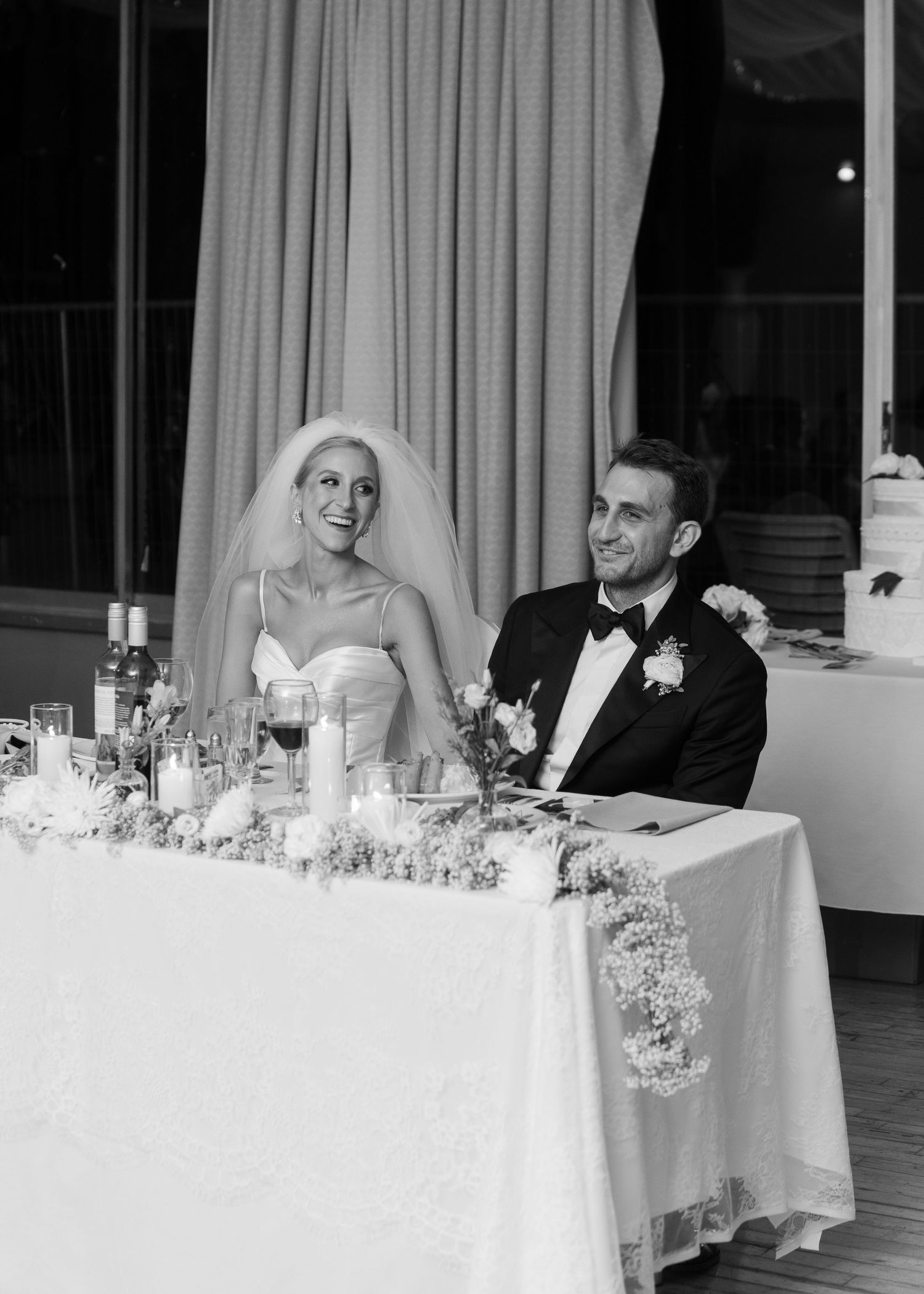 Bride and groom smile at sweetheart table adorned with candles and floral garland at reception.