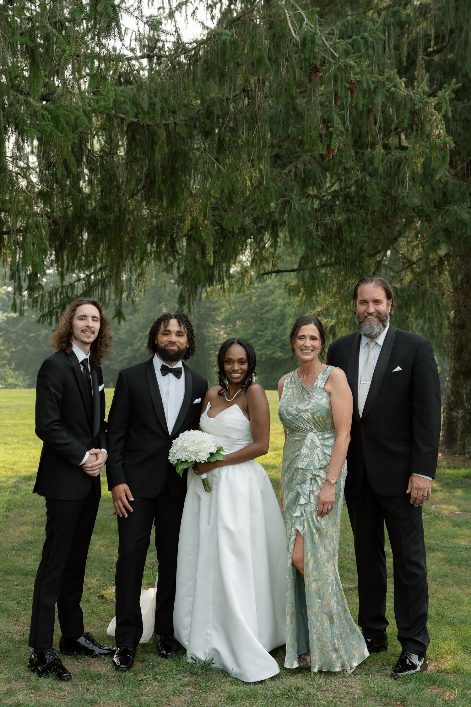 Wedding couple poses with family under evergreen tree, bride in white gown holding bouquet, groom in tuxedo.