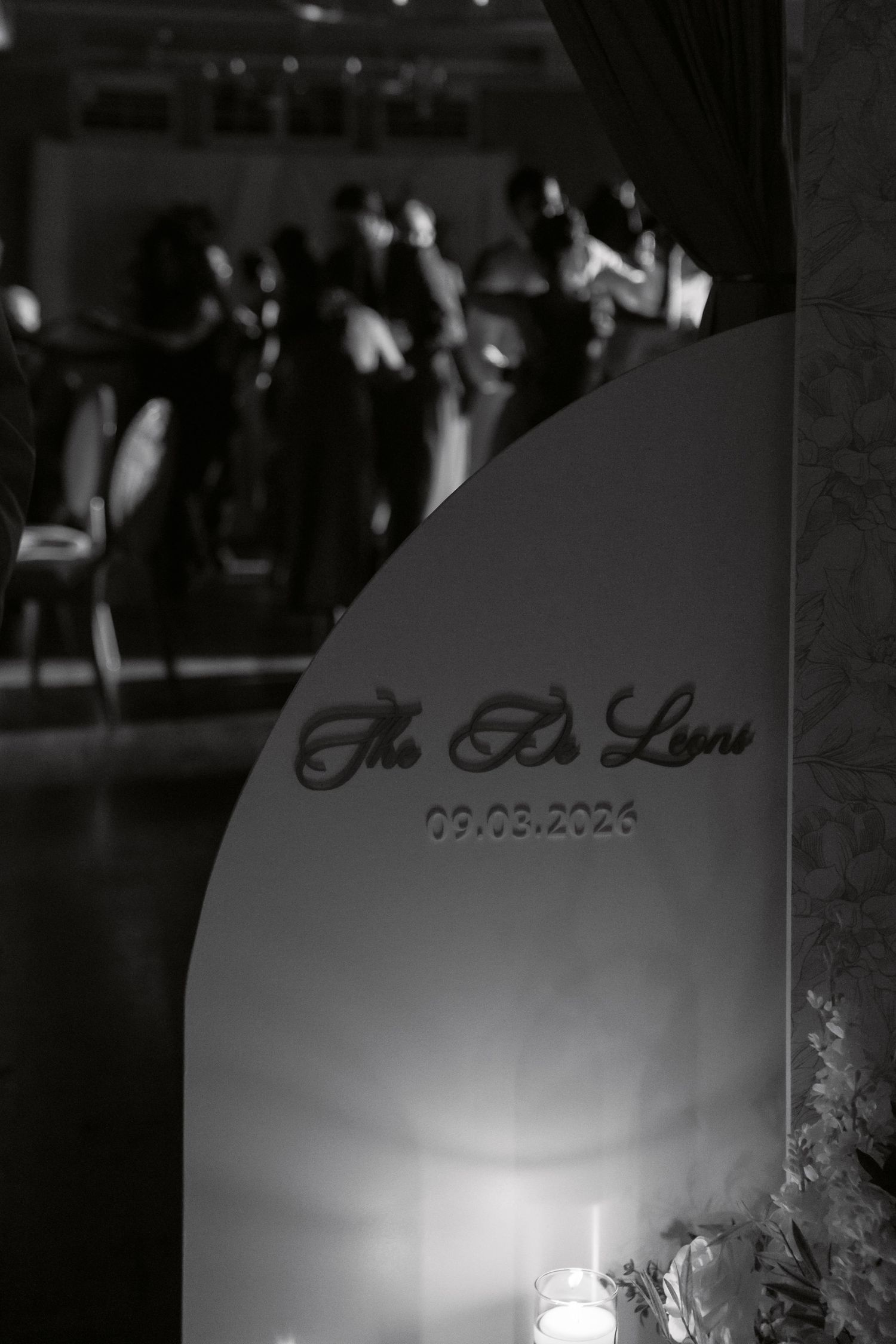 Black and white wedding reception photo featuring The De Leon family name sign dated 09.03.2026 with guests dancing in background.
