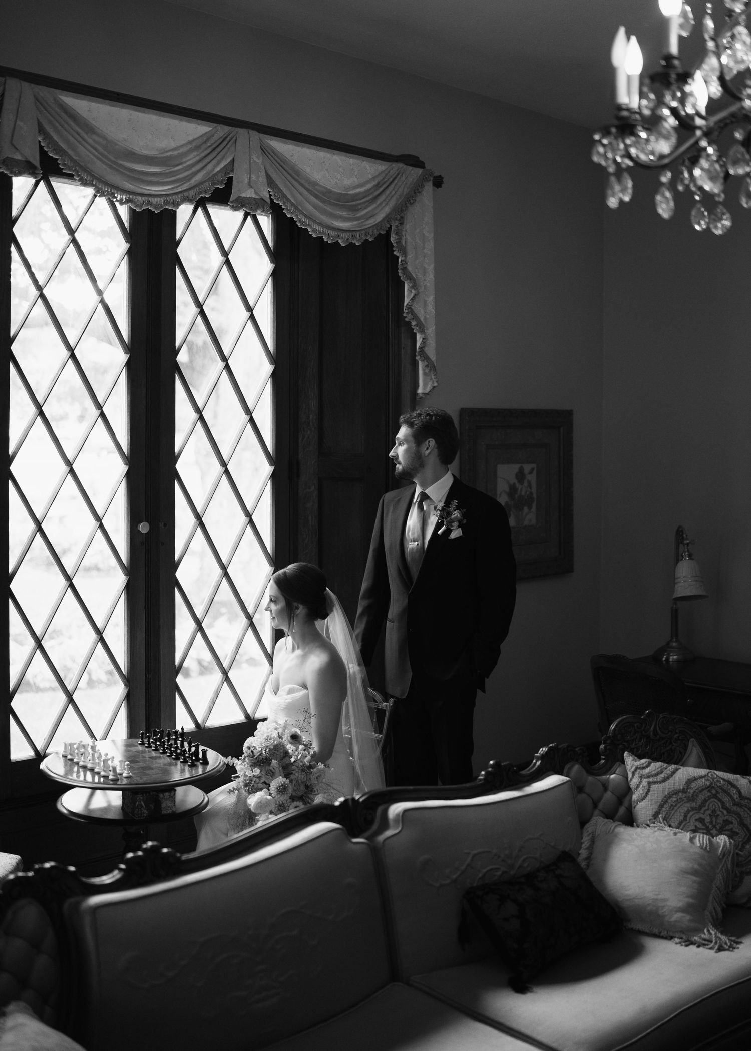 Black and white wedding photo of bride and groom standing by diamond-paned windows in elegant room with chandelier.