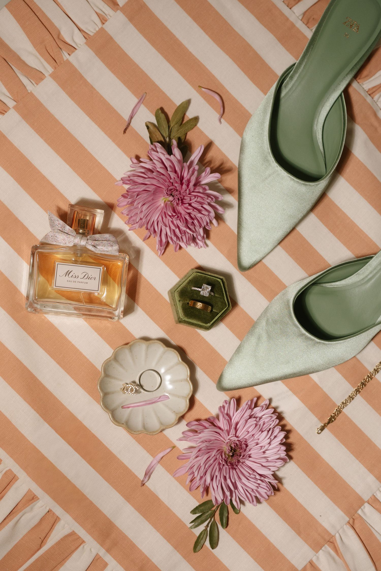 Flatlay of mint green heels, perfume bottles, pink chrysanthemums and wedding rings on peach striped background.