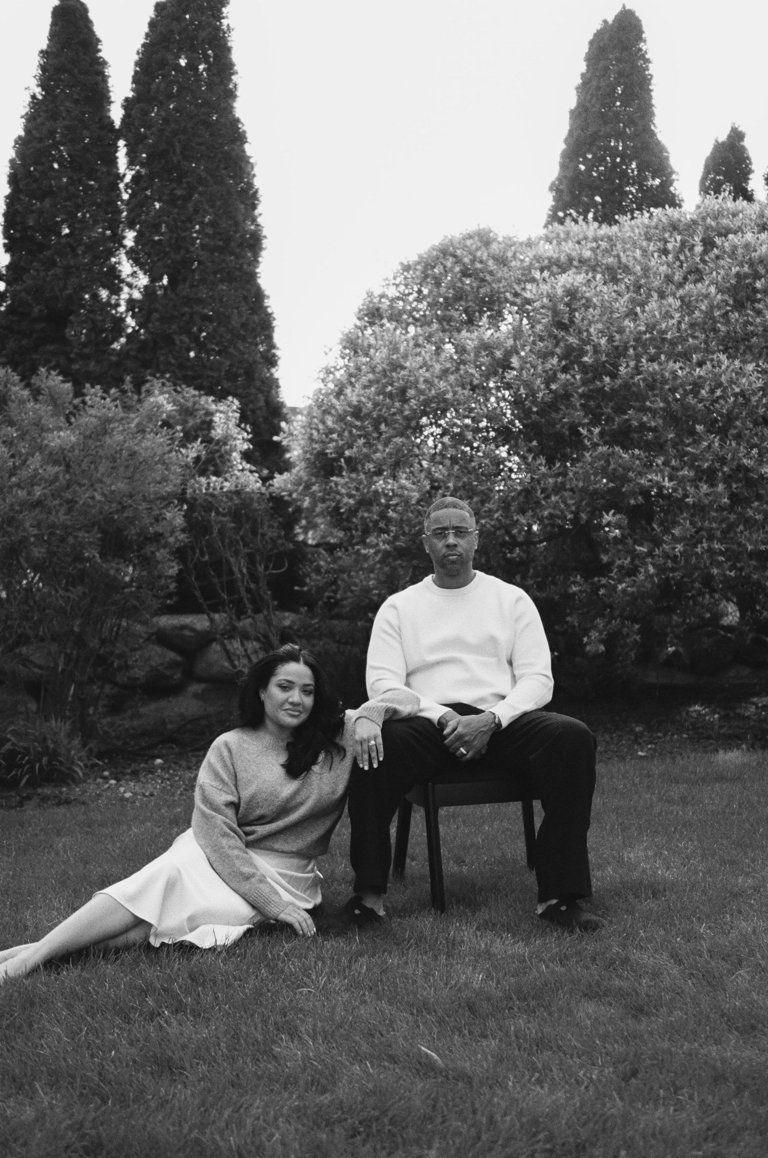 Black and white photo of a couple posing in a lush garden, woman sitting on grass, man seated on a chair.